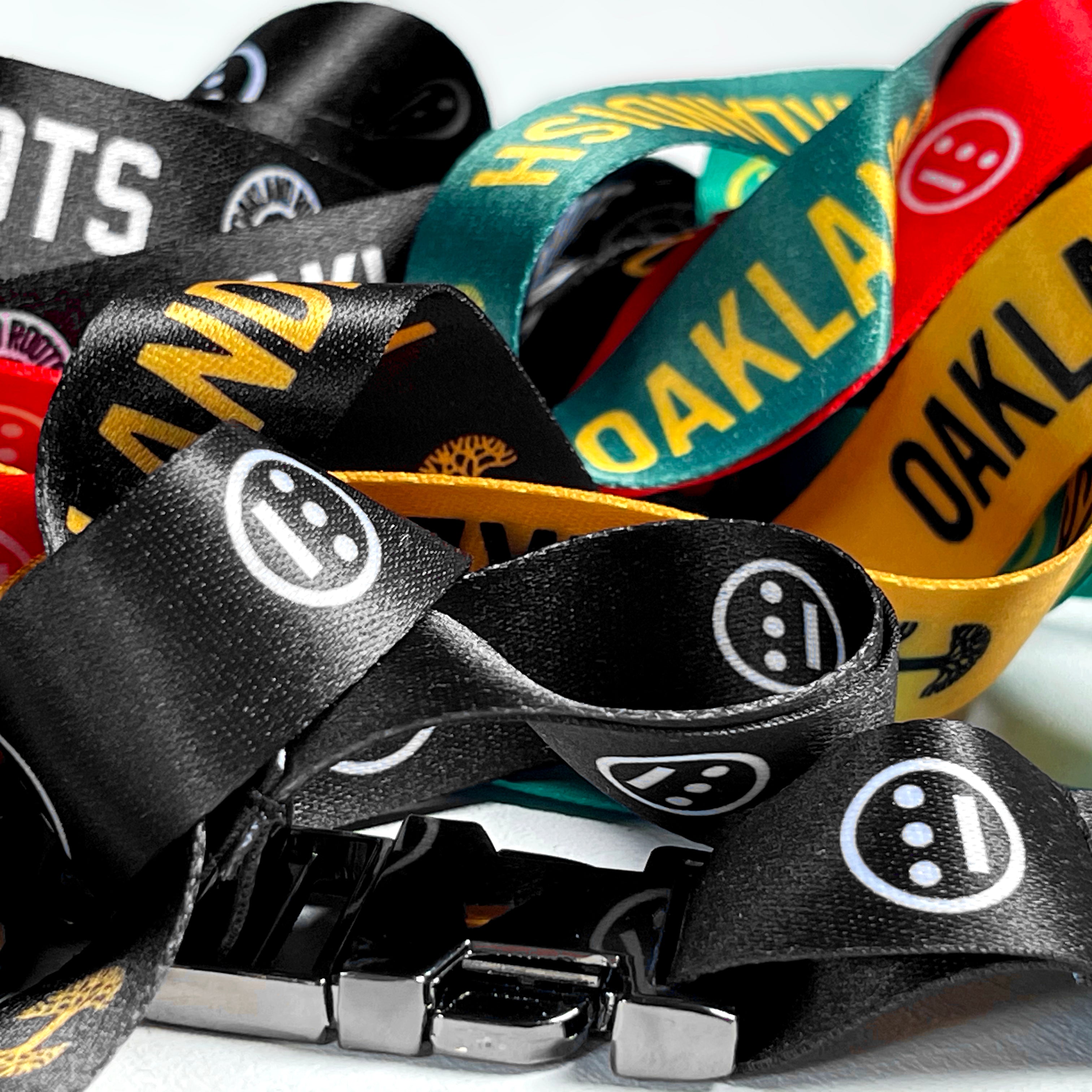A vibrant display of Hiero VIP Lanyard 2.0 from Hieroglyphics features various hip-hop designs, including text like "OAKLAND" and distinctive logos. The lanyards come in black, yellow, red, and green. Intertwined together, a metal clasp is prominently visible at the forefront, showcasing their texture and vivid colors in a close-up shot.