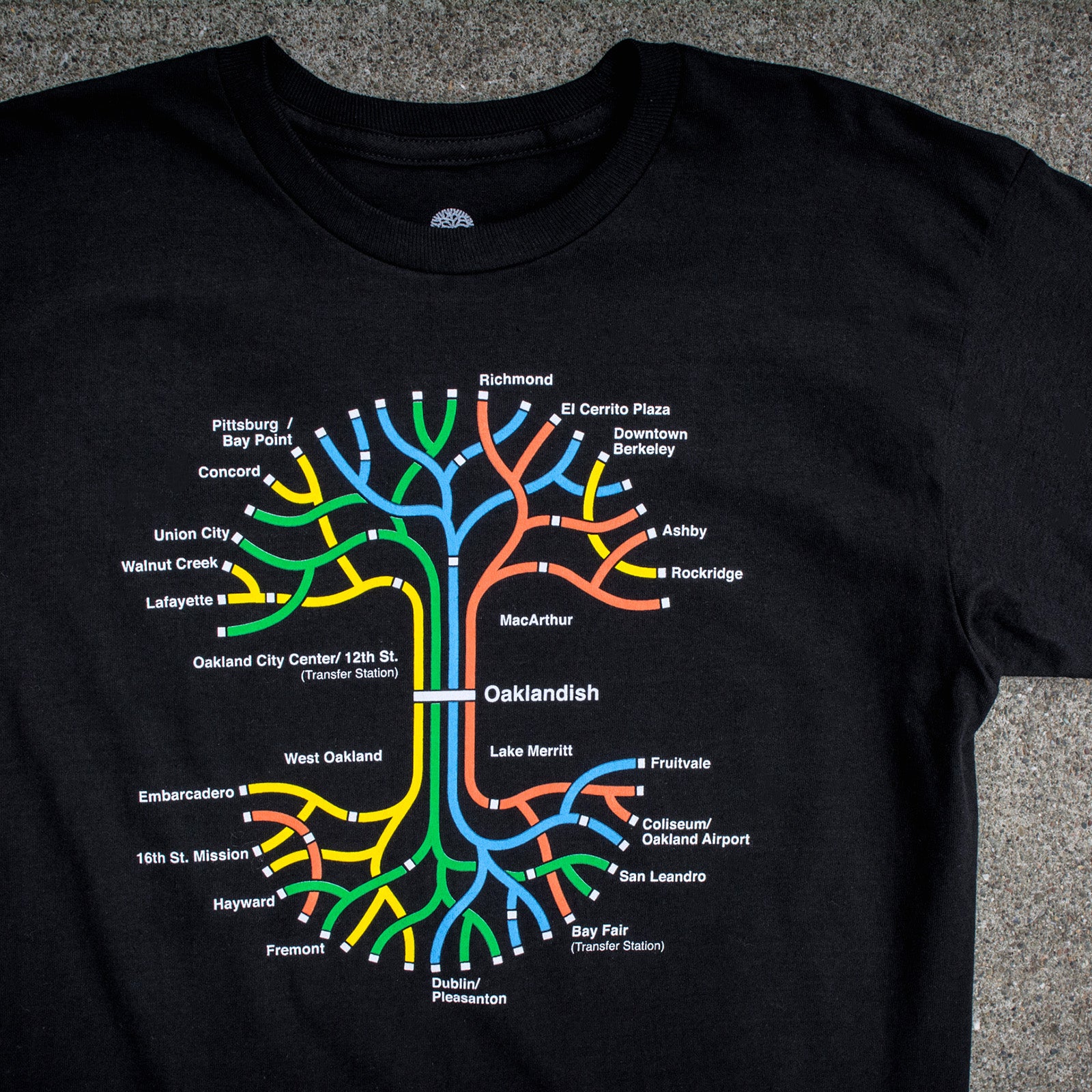 The BART Tree Tee by Oaklandish is a 100% cotton, classic fit t-shirt that features a vibrant map of the Bay Area Rapid Transit (BART) system. The design showcases various station names and lines in red, blue, yellow, orange, and green, all branching out from a central point labeled "Oaklandish," beautifully centered on the shirt.
