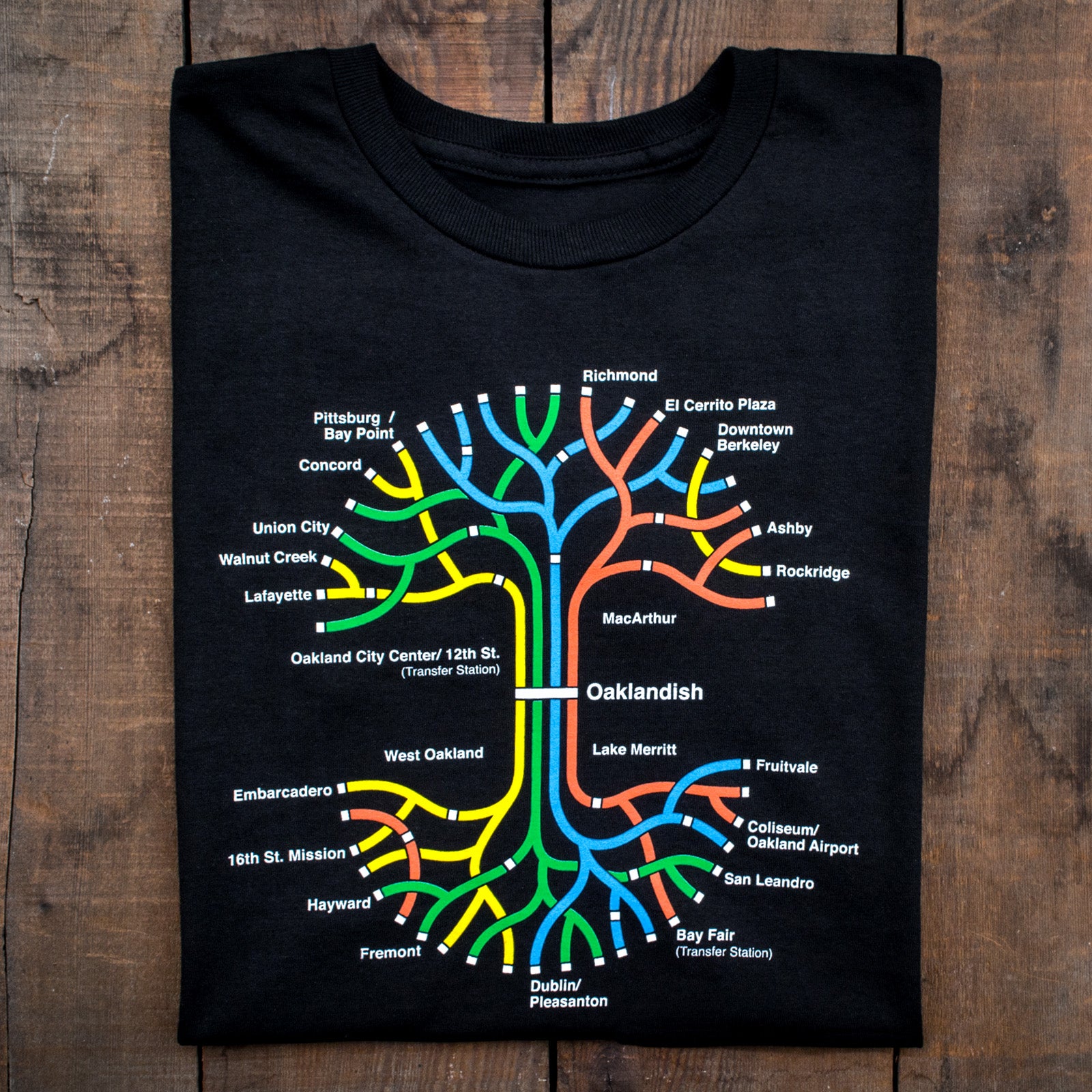 The Oaklandish BART Tree Tee, made from 100% black cotton, is displayed on a wooden surface. This classic fit t-shirt features a colorful, stylized BART map of the San Francisco Bay Area with stations like Civic Center, 12th St., and Concord. The design showcases lines in red, yellow, blue, green, and orange.