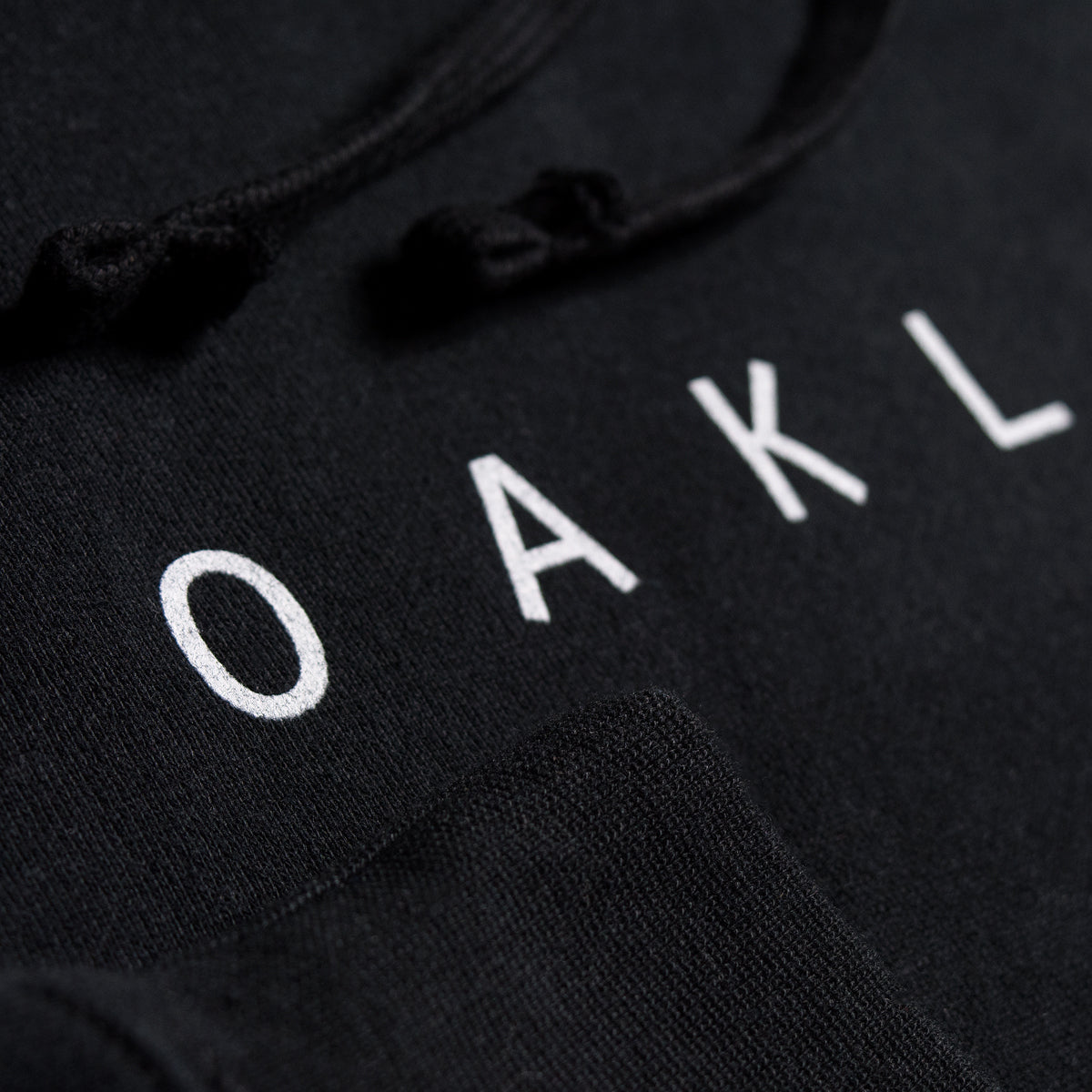 Close-up photo of the Oaklandish Classic Hoodie by Oaklandish, a black pullover featuring white text with partially visible letters spelling "OAKL." The men's fit hoodie from this brand includes black drawstrings, and the fleece fabric is detailed in the image, showcasing its texture and stitching. The lettering is printed or embroidered in a clean, minimalist font that hints at its Oakland roots.