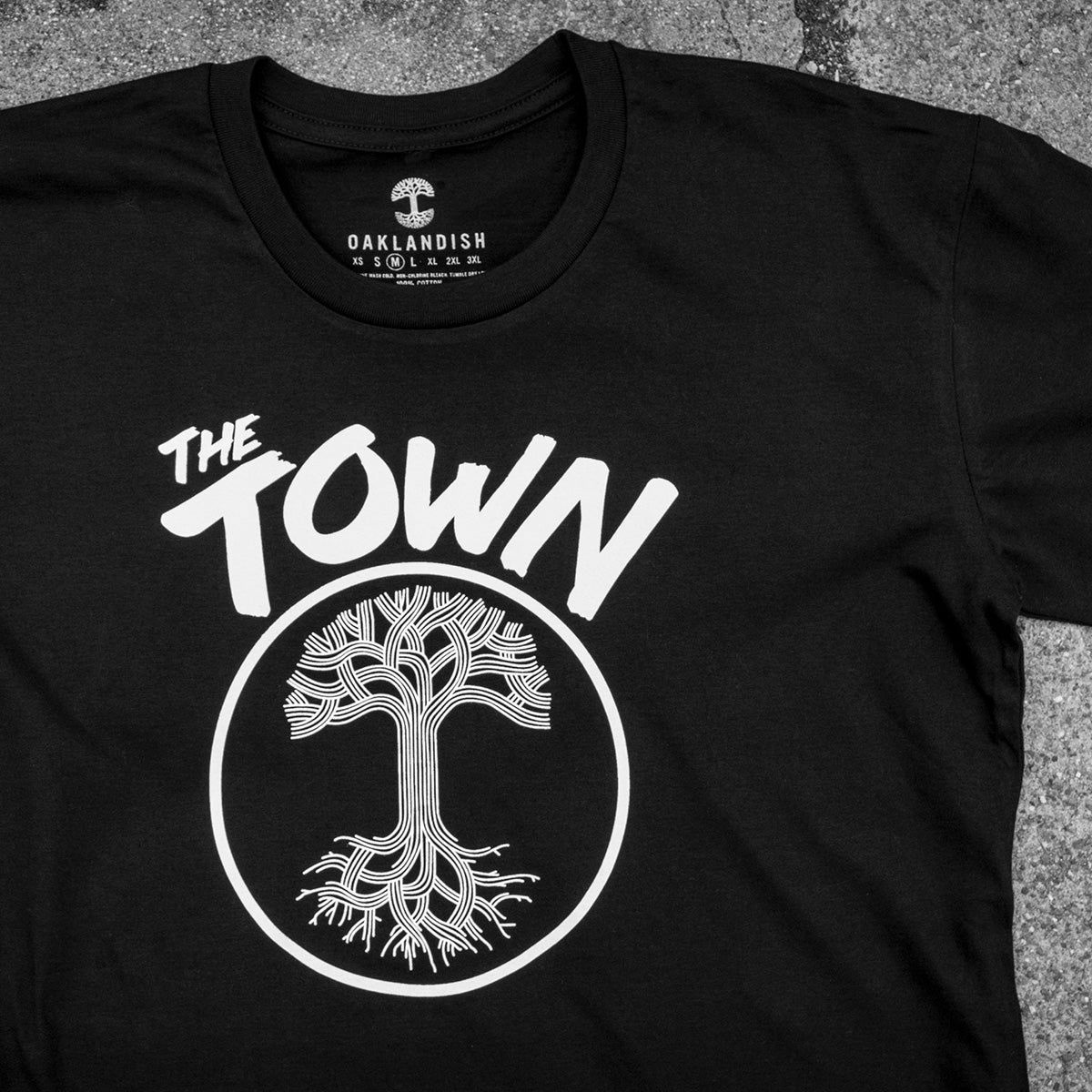 The Forever Tee from Oaklandish, part of the Men's collection, is a black classic fit t-shirt displayed on a gray surface. It features the phrase “The Town” in white bold, playful letters, with a tree encircled by intricate roots and branches below the text. Made from 100% cotton, this tee also showcases a small tree icon and the slogan “I (heart) Oakland.”