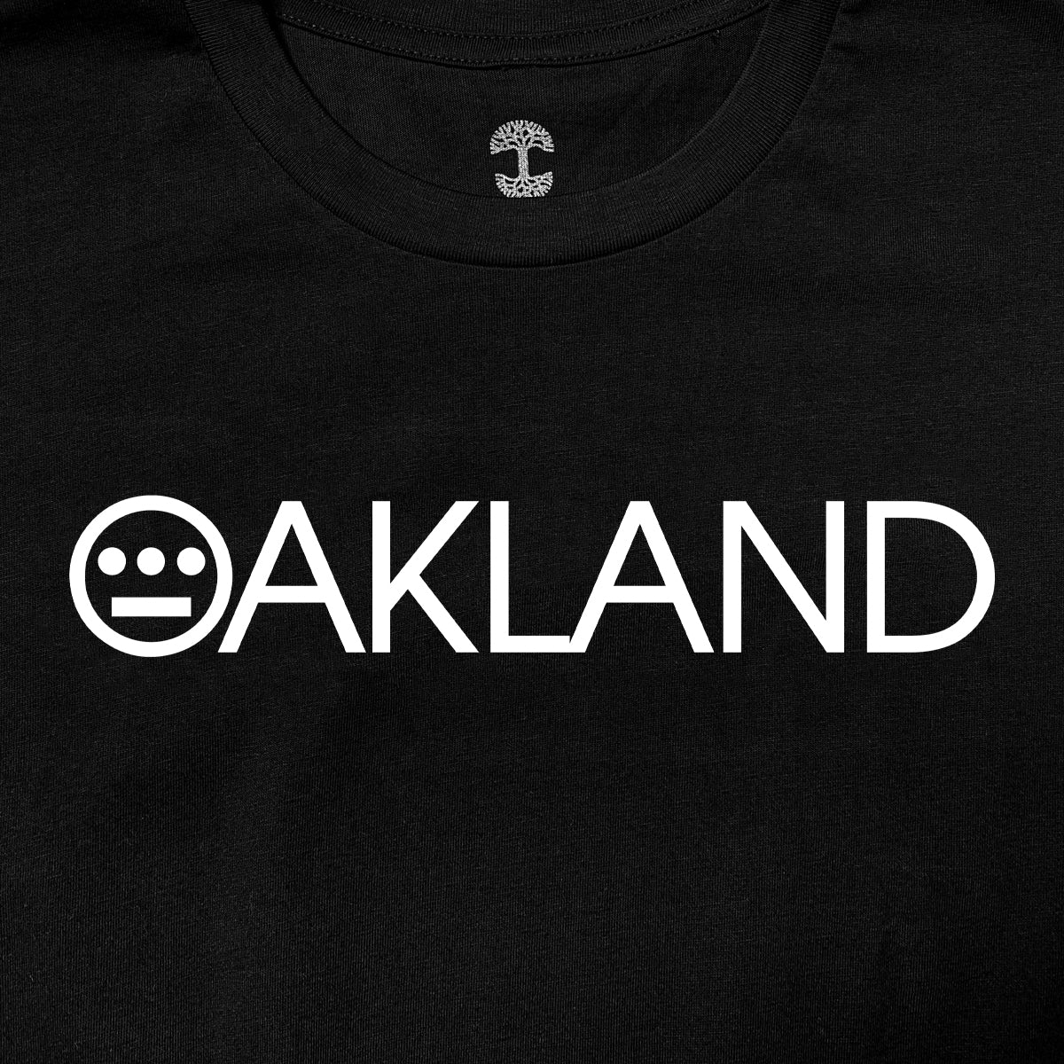The Hieroakland Tee by Hieroglyphics is a black, 100% cotton T-shirt featuring the word "OAKLAND" in bold, white capital letters across the chest. The "O" is uniquely stylized with three horizontal dots above two vertical lines. Inside the neckline area, there's an intricate tree design that adds a special touch to this classic fit T-shirt, making it perfect for any town business.