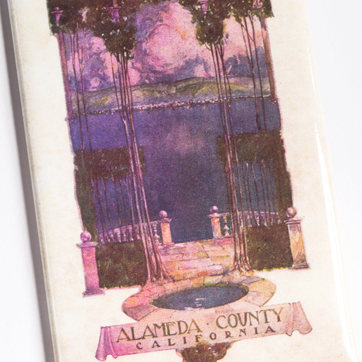 The Alameda County Magnet by Oaklandish features a vintage postcard image showcasing a scenic view of Alameda County, California. The artwork highlights a stone pathway leading through an ornate archway, framing a serene body of water with the hilly landscape of the East Bay in the background. "Alameda County, California" is prominently written at the bottom, now available as a charming refrigerator magnet!