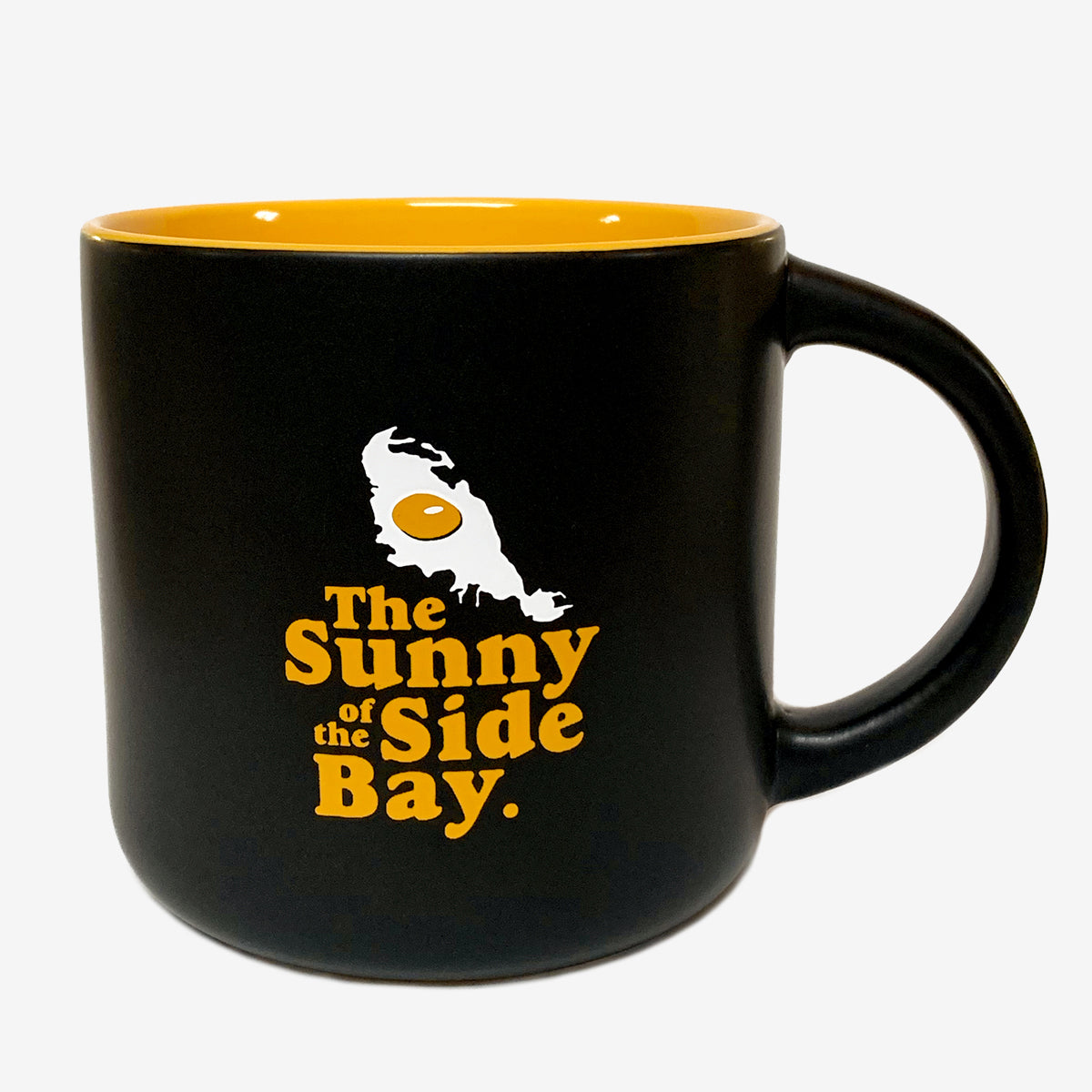 The Sunny Side Mug by Oaklandish features black stoneware with a glossy yellow interior. The front boasts an illustration of a sunny-side-up egg above an outline of a landmass, accompanied by the text "The Sunny Side of the Bay" in yellow and white colors.