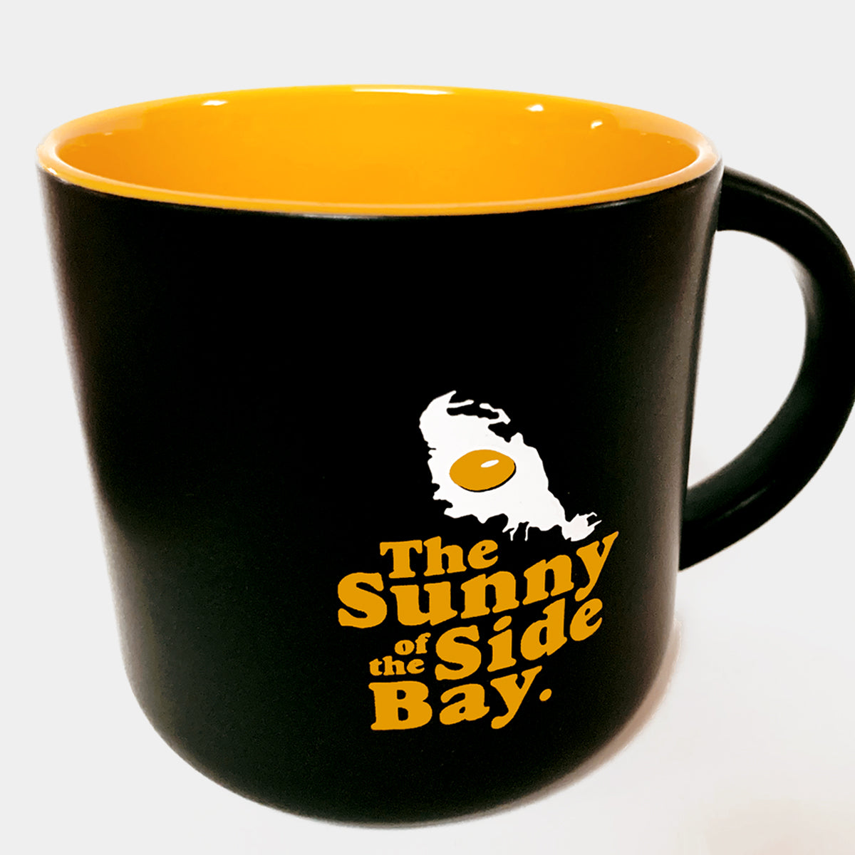 Presenting the Sunny Side Mug from Oaklandish: A black ceramic bay mug featuring a glossy yellow interior. Adorning the mug is a graphic of a fried egg accompanied by the text "The Sunny Side of the Bay" in prominent yellow letters on its side.