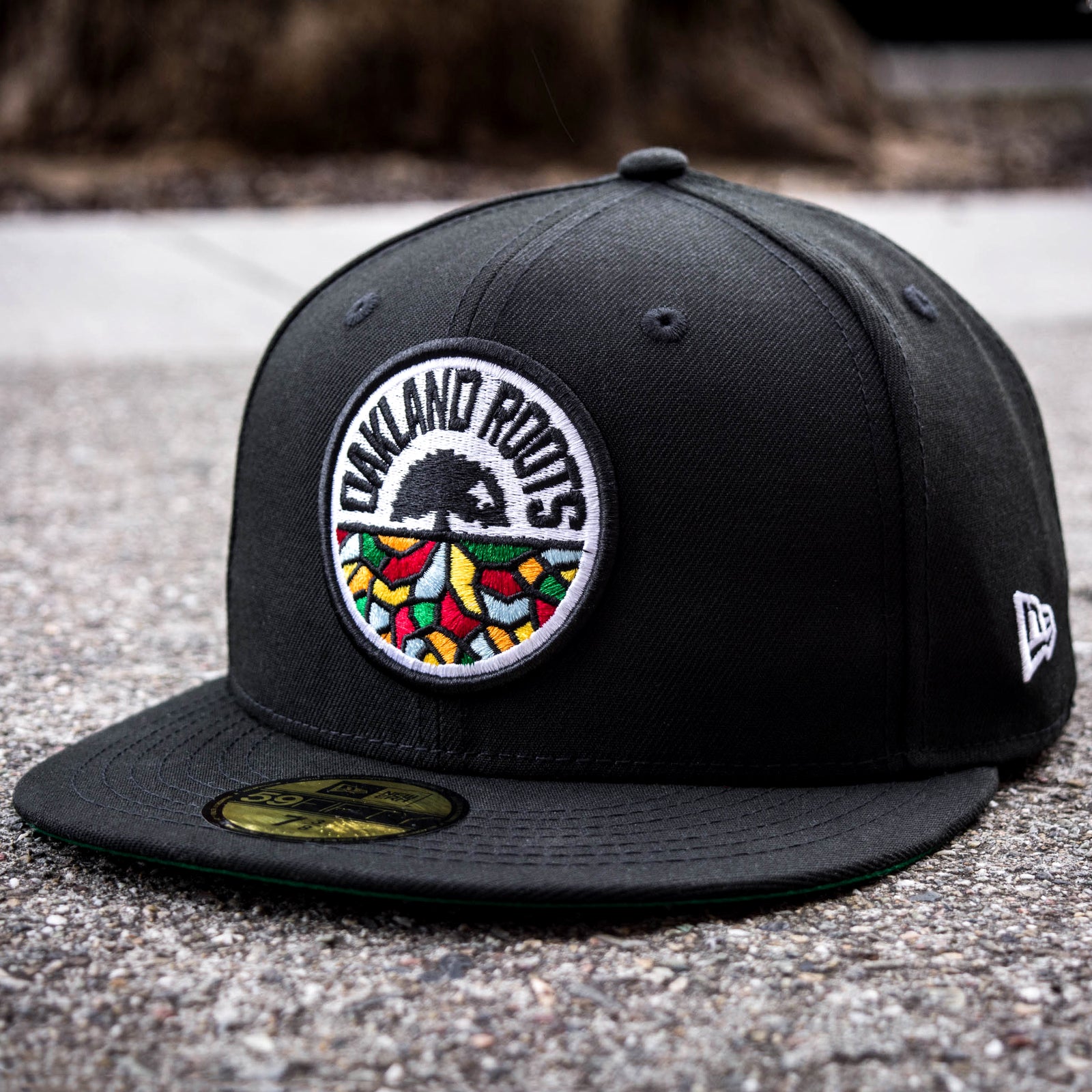 A black New Era Oakland Roots SC 59FIFTY Fitted Cap prominently displaying a circular embroidered logo on the front. The logo features a tree with "OAKLAND ROOTS" inscribed around it, with colorful patterns resembling leaves or roots at the bottom. The hat rests on a textured surface, and a small white Oakland Roots SC emblem adorns the side.