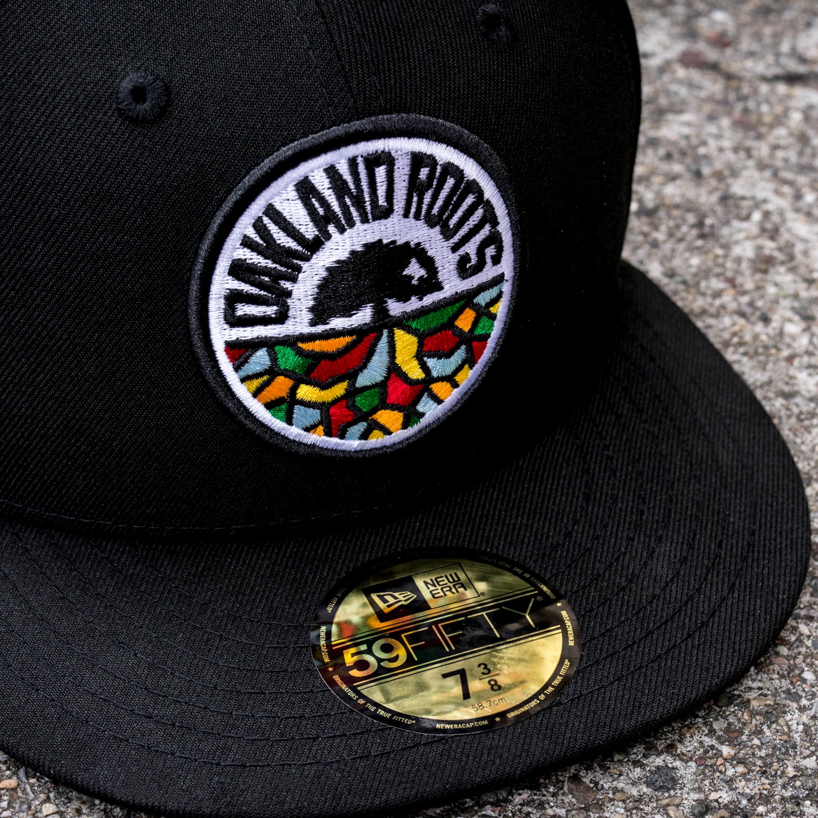 A New Era Oakland Roots SC 59FIFTY Fitted Cap rests on a gravel surface. The front of the black cap features an embroidered "Oakland Roots" logo, depicting a stylized tree with multicolored roots that highlight the sports club's emblem. A round gold New Era 59FIFTY sticker, displaying cap size 7 3/8, is affixed to the brim.