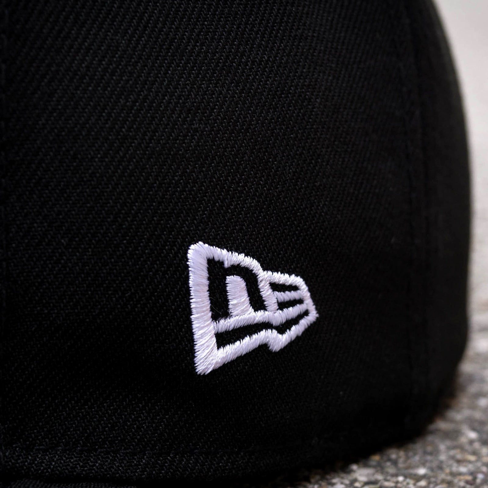 Close-up of the New Era Oakland Roots SC 59FIFTY Fitted Cap in black, showcasing an embroidered white logo. The logo features a stylized "N" followed by a smaller "E" in a continuous line, creating a distinctive design. The texture of the fabric and the precision of the stitching are clearly visible, making it perfect for Oakland Roots SC fans.