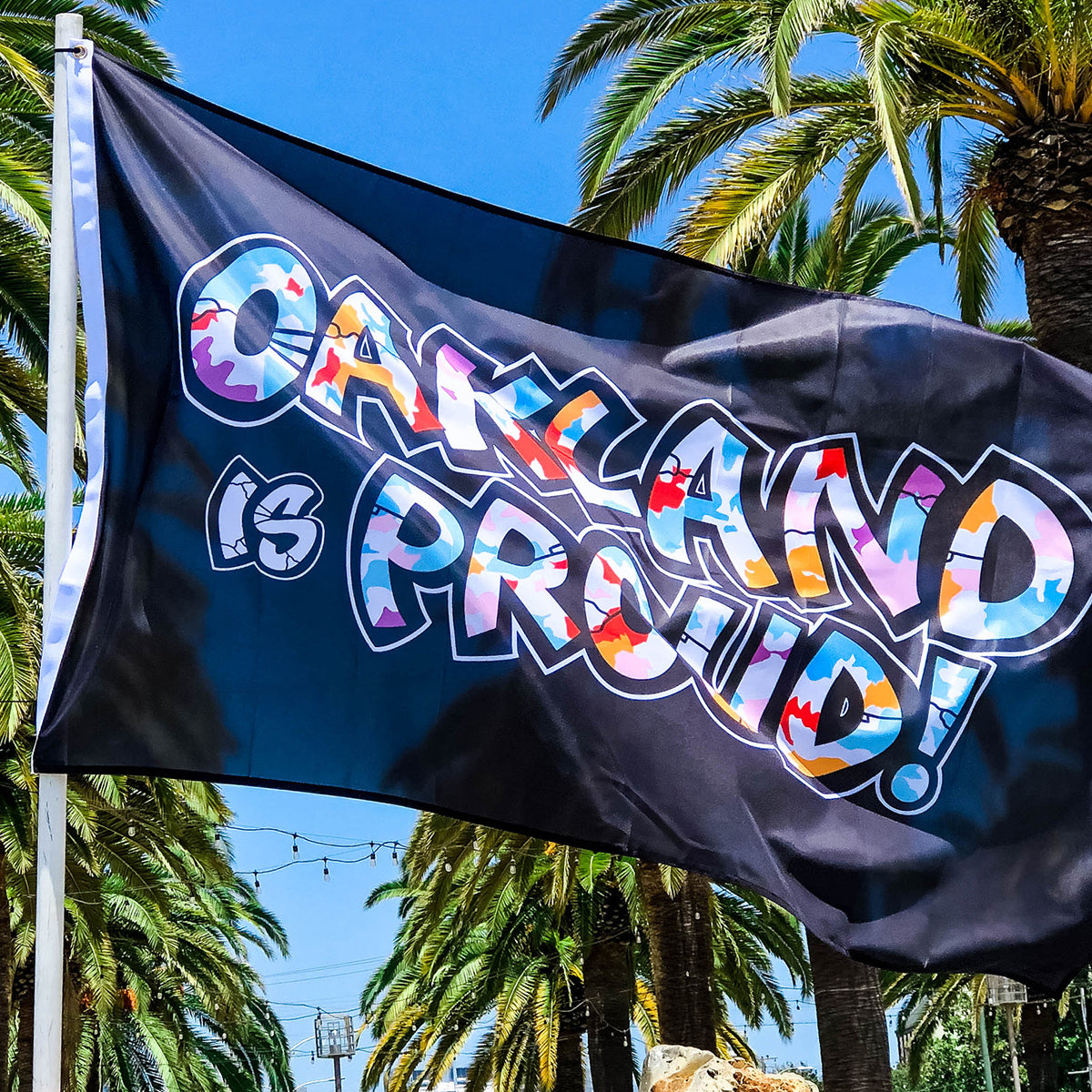 A black flag, named the "Oakland is Proud Flag" from Oaklandish, features colorful graffiti-style text proclaiming "Oakland is Proud!" against a backdrop of lush palm trees and a clear blue sky. The vibrant colors of the text stand out prominently, much like an Oakland mural by Del Phresh, enhancing the lively and celebratory atmosphere of the scene.