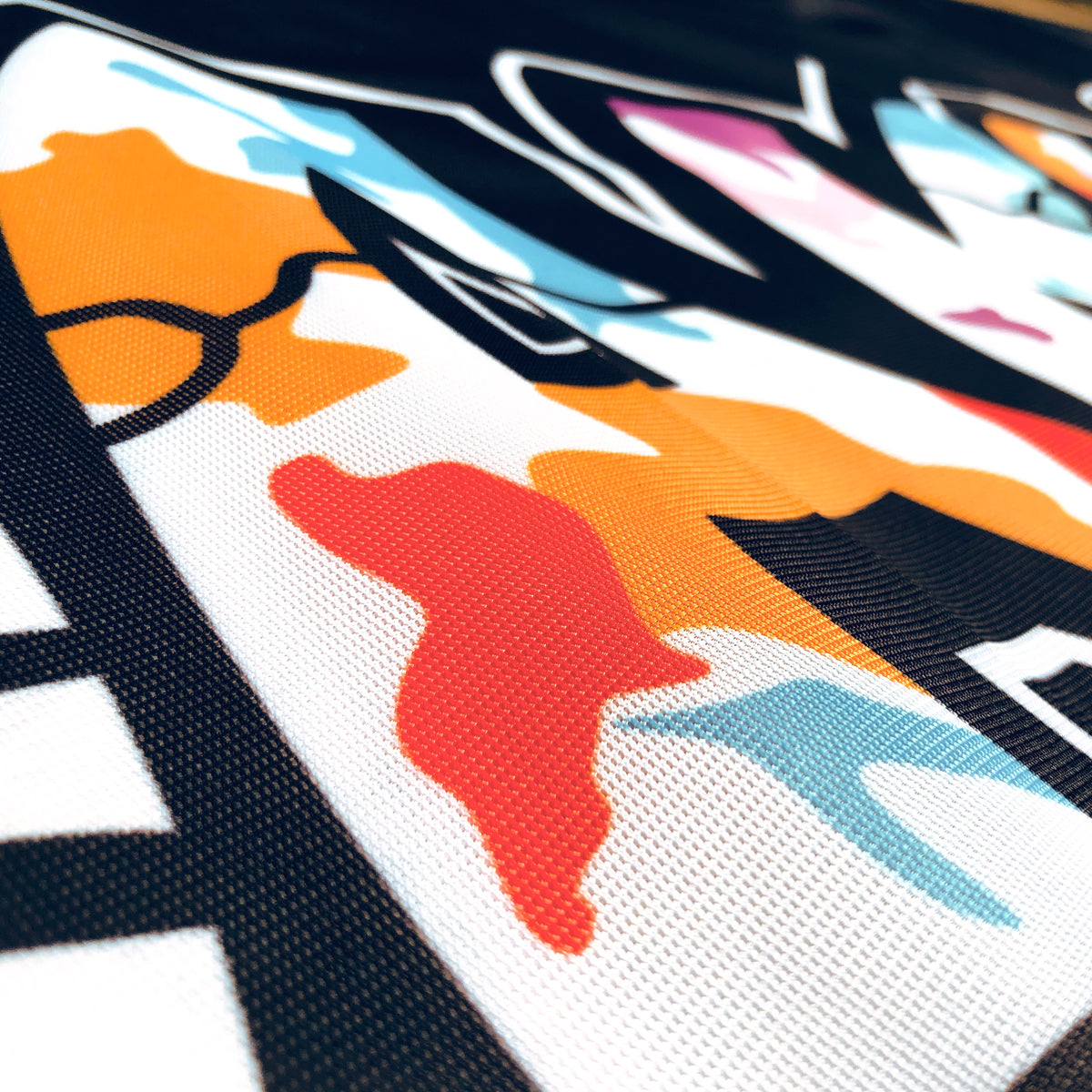A close-up image of the Oakland is Proud Flag by Oaklandish features vibrant abstract shapes in orange, red, blue, pink, and black on a white backdrop. The fabric's texture is clearly visible, with woven details and slight creases enhancing its appearance. This artistic and visually striking design would make for stunning accessories.