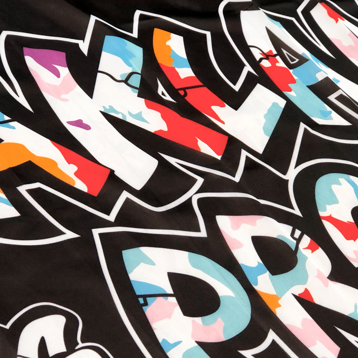The **Oakland is Proud Flag** by **Oaklandish** features graffiti-style text with colorful camo patterns against a black background. The bold, jagged letters are outlined in white and filled with a mix of red, blue, pink, light blue, and orange colors. This dynamic design exudes an urban street art aesthetic that is reminiscent of an iconic Oakland mural.