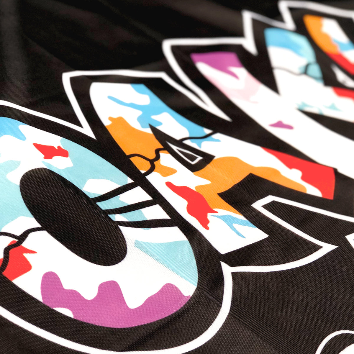 A close-up of bold, stylized letters with a camouflage pattern in various colors like blue, orange, pink, and white. The letters are outlined in white and set against a black background. This glimpse showcases the vibrant color palette of the Oaklandish "Oakland is Proud Flag," resembling unique accessories for the eyes.