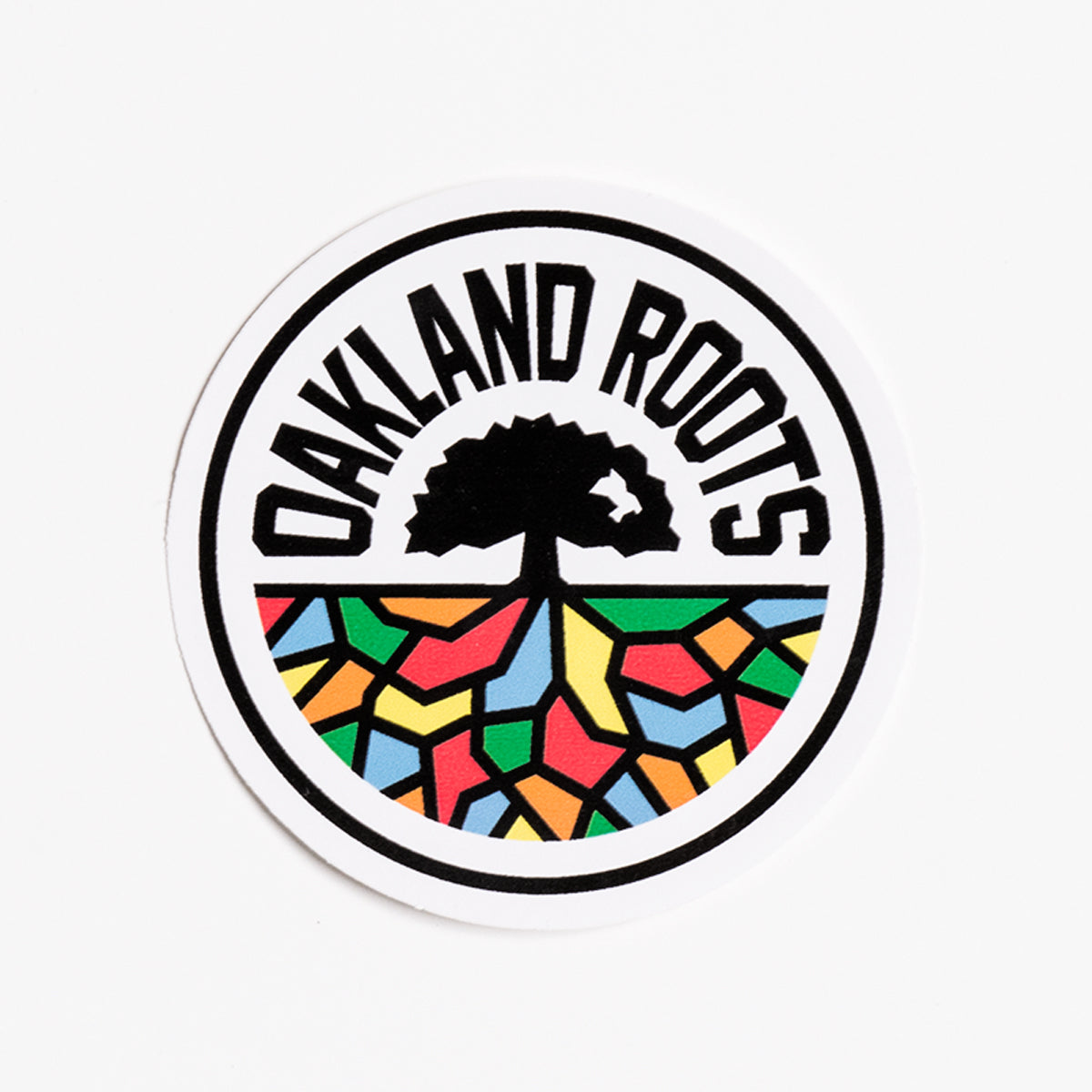The Oakland Roots SC Logo Sticker from Oakland Roots SC features a circular logo with "OAKLAND ROOTS" in bold, black letters at the top. The center presents a stylized black tree with spreading roots, while below, vibrant geometric shapes in red, yellow, green, blue, and black fill the lower half, reminiscent of an Oaklandish Sticker with a stained glass design.
