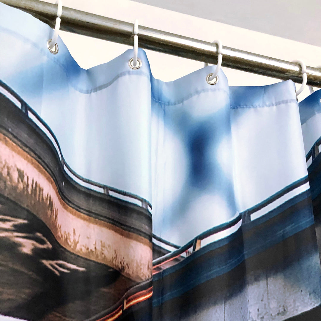 A close-up of the Overpass Shower Curtain 72 X 72 by Oaklandish is shown hanging from a metal rod with metal hooks. The polyester shower curtain showcases an abstract design with blurred, wavy lines in soft blue, gray, and brown hues, giving it a modern and artistic look. The top features metal grommets for the hooks, making it ideal for any bathroom.