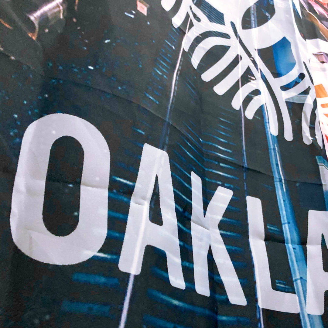 Close-up of the Overpass Shower Curtain 72 X 72 by Oaklandish, resembling a fabric banner with large white text "OAK LA" against a background depicting train tracks. The shower curtain also features white curved lines, possibly part of a larger image or logo, and the fabric shows visible folds and creases.