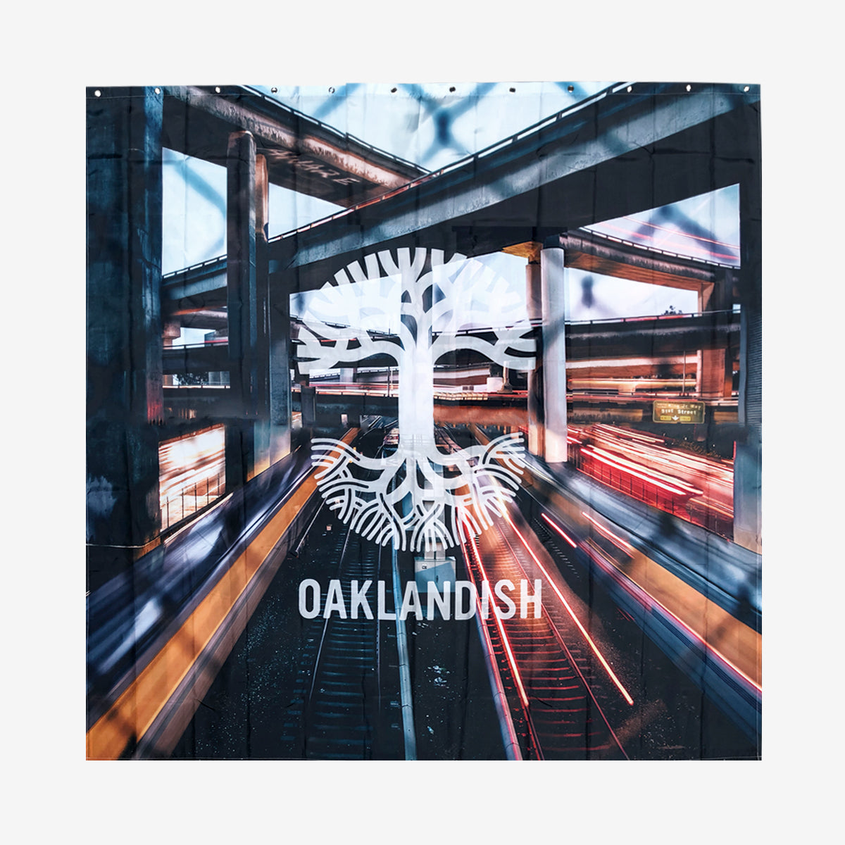 Image showcases the "Overpass Shower Curtain 72 X 72" by Oaklandish. The design features a stylized white tree logo with intertwined roots and branches, overlaid with the word "OAKLANDISH" in white text. The background, reminiscent of a polyester shower curtain, depicts a blurred urban scene with multi-level highways and streaks of light from moving vehicles, evoking a sense of motion and urban energy.