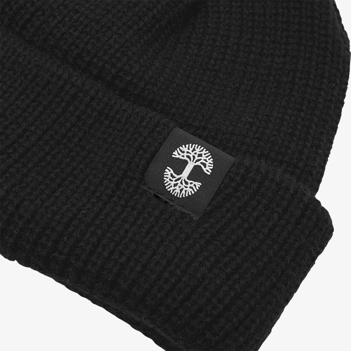 Here's a close-up view of the Oaklandish Ramos Beanie folded at the brim. This black knit headwear showcases a small black square patch with an embroidered white tree design on the fold. The texture of its woven acrylic fabric is clearly visible, emphasizing its ribbed pattern.