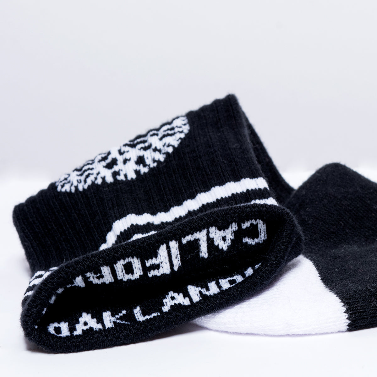 The Retro Classic Socks from Oaklandish, a pair of black socks with white lettering and designs, are shown. One sock features the word "CALIFORNIA" while the other displays "OAKLAND," both partially visible. These exceptionally comfy socks boast white details along with an Oaklandish logo and are displayed on a white surface.