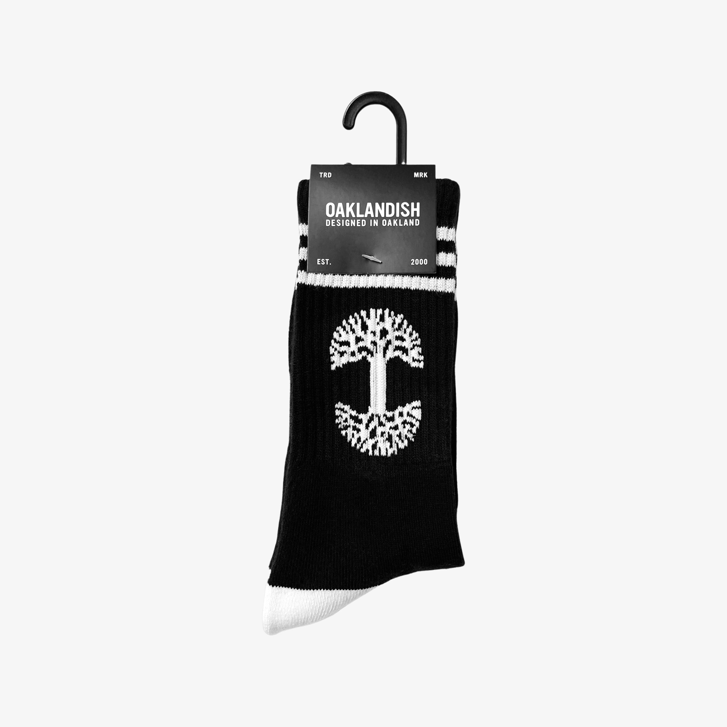 A pair of exceptionally comfy Retro Classic Socks by Oaklandish, featuring a black base with white accents. These socks showcase a white tree symbol in the center, white toes, and two white stripes near the top. The packaging label proudly displays "Oaklandish," along with "Designed in Oakland" and "EST. 2000.
