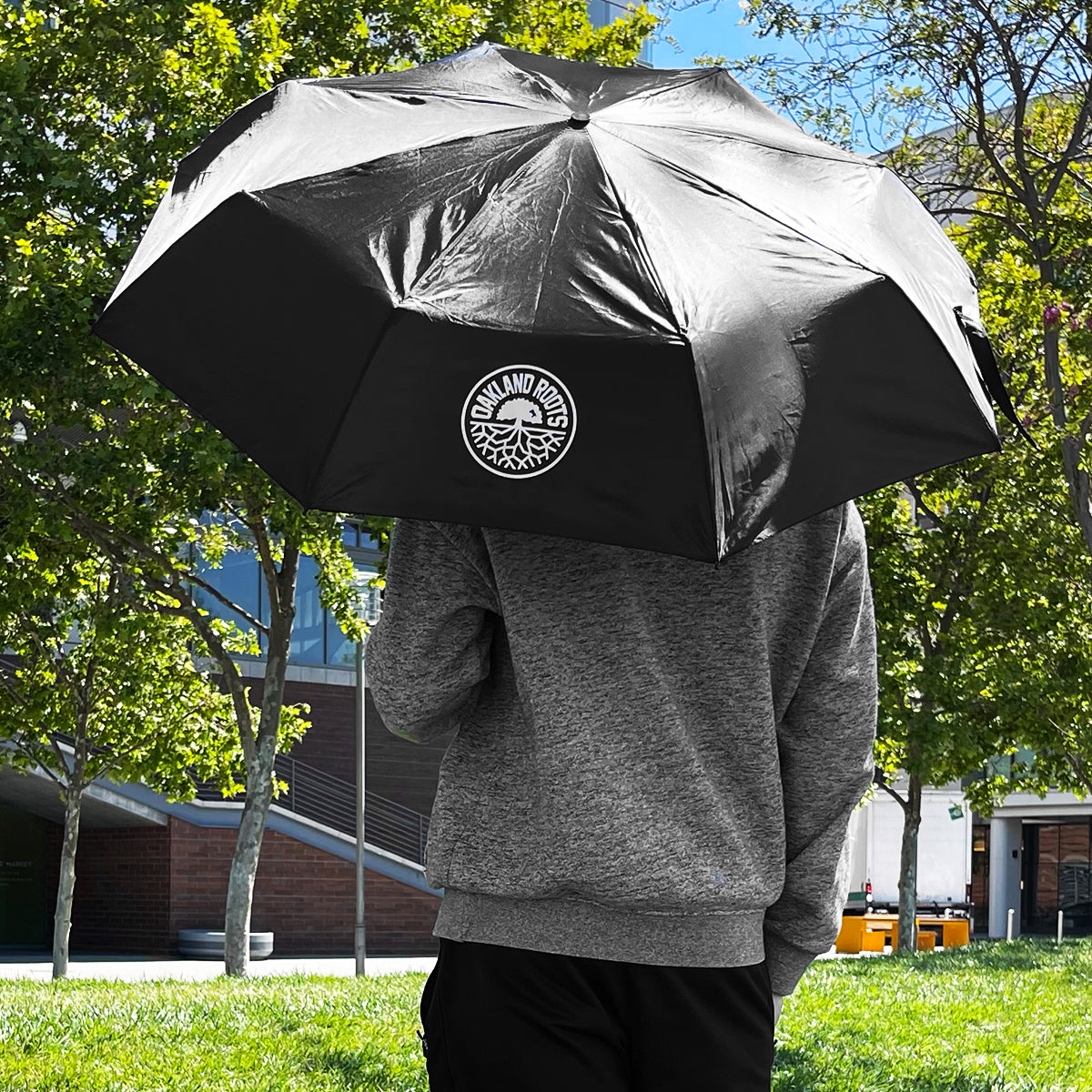A person in a gray sweatshirt holds the Oakland Roots SC Compact Umbrella, which features the Oakland Roots SC logo. They stand in a park with lush green trees and modern buildings in the background. The focus is on the umbrella, and the person is partially turned away from the camera.