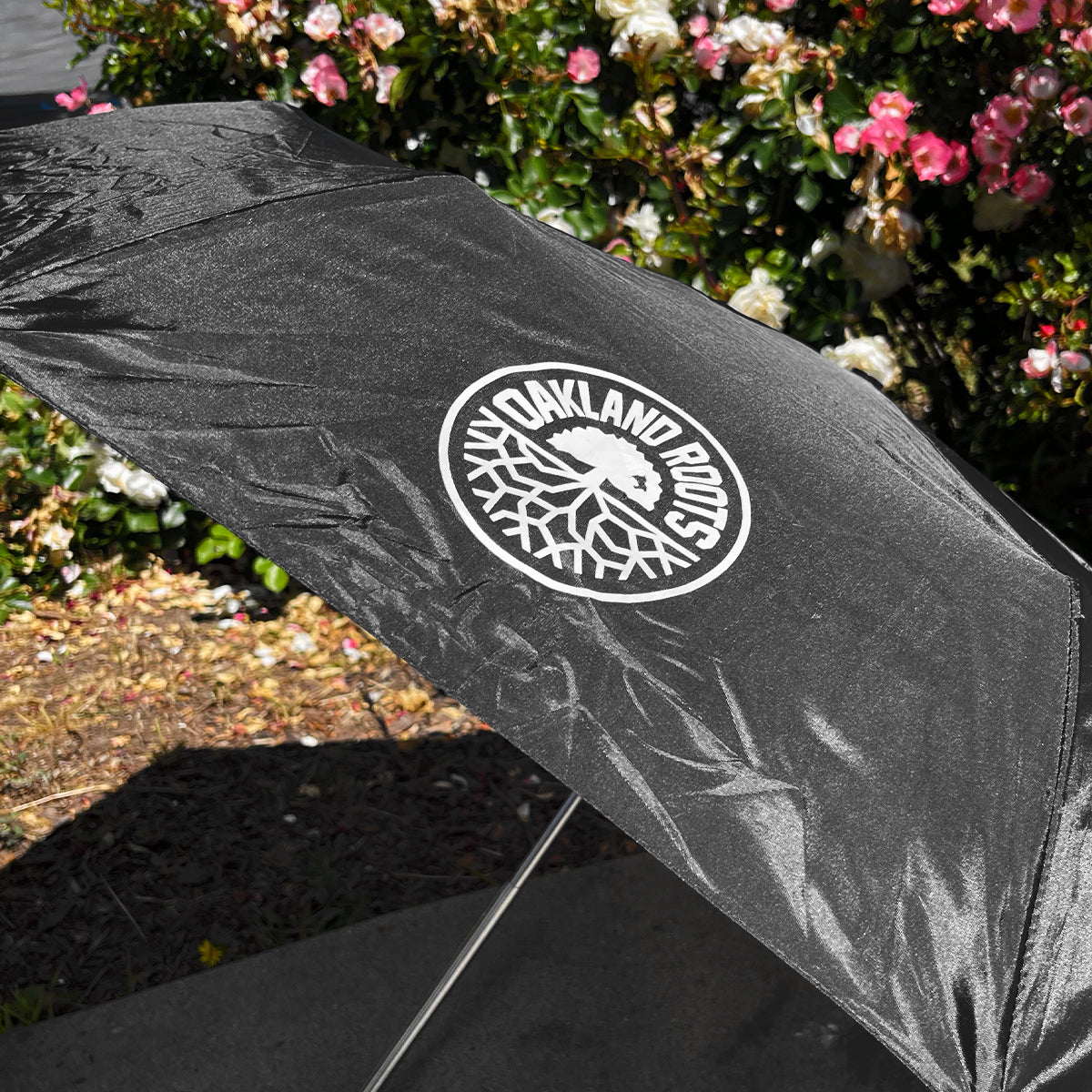 An Oakland Roots SC Compact Umbrella, in black, is fully open outdoors, offering shade. The umbrella flaunts the Oakland Roots SC logo in white, showcasing a stylized tree with roots. Around it, vibrant blooming flowers and lush green foliage form a colorful and lively backdrop.