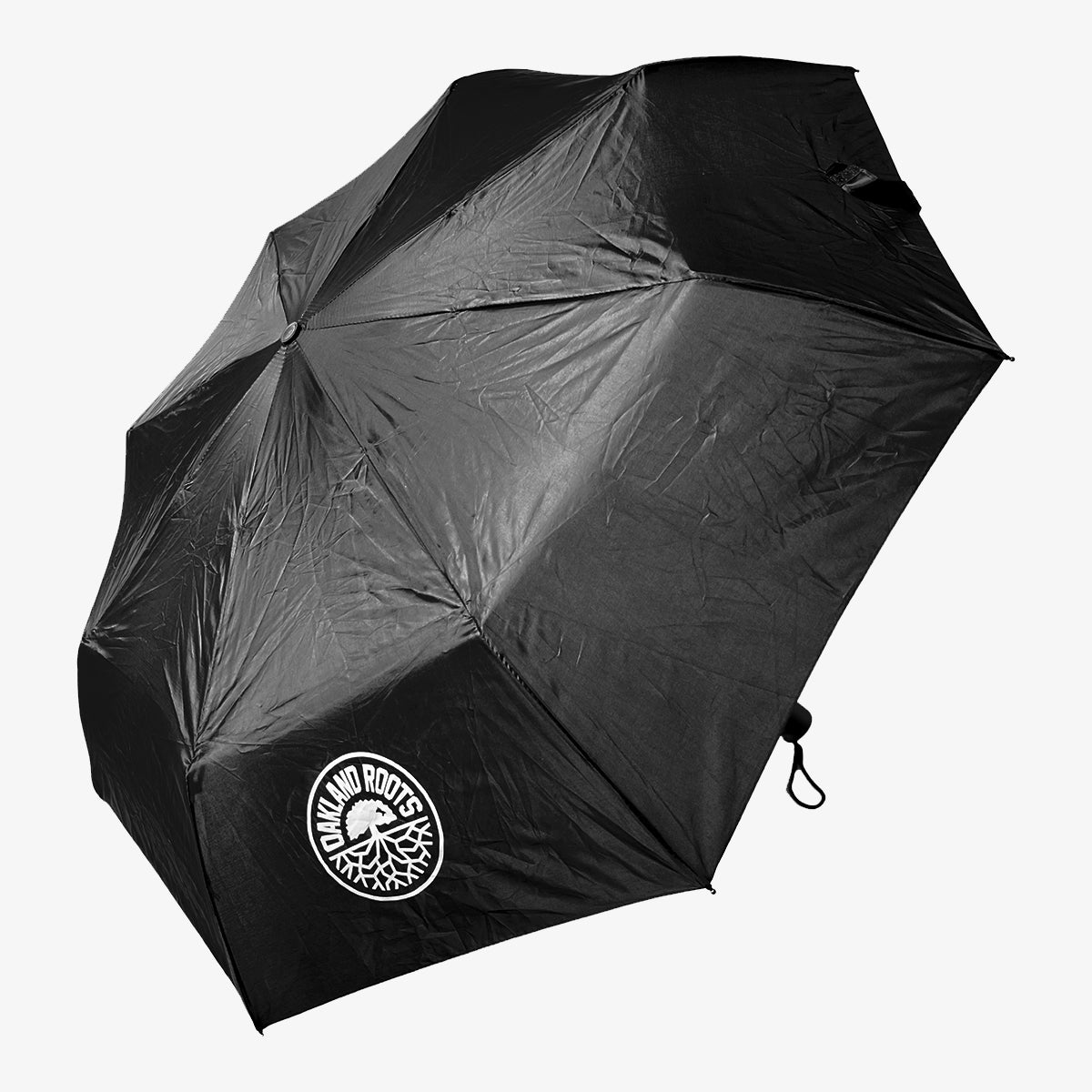 A black Oakland Roots SC Compact Umbrella with a slightly crumpled canopy. It features a logo near the edge consisting of a tree with broad roots and the words "TOWA AND ROOTS" encircling it, hinting at its Oakland Roots inspiration. The umbrella handle has a black strap attached. The background is plain white.