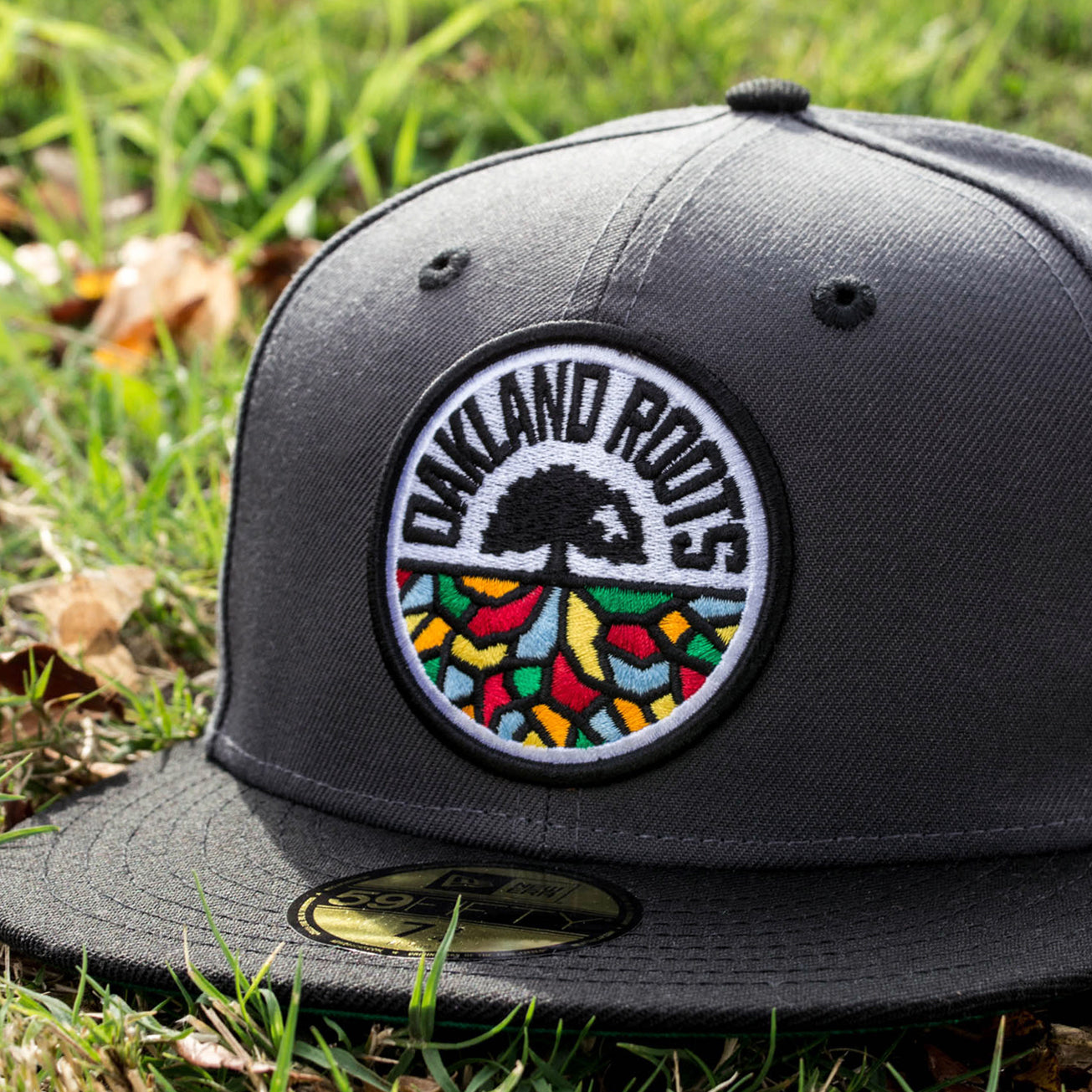 A New Era Oakland Roots SC 59FIFTY Fitted cap in gray, featuring an "Oakland Roots" logo with a black tree and roots extending into colorful geometric shapes, surrounded by a circular white border that includes the text "OAKLAND ROOTS," is lying on the grass. The cap also includes a gold sticker on its bill, making it perfect for any Oakland Roots SC supporter.