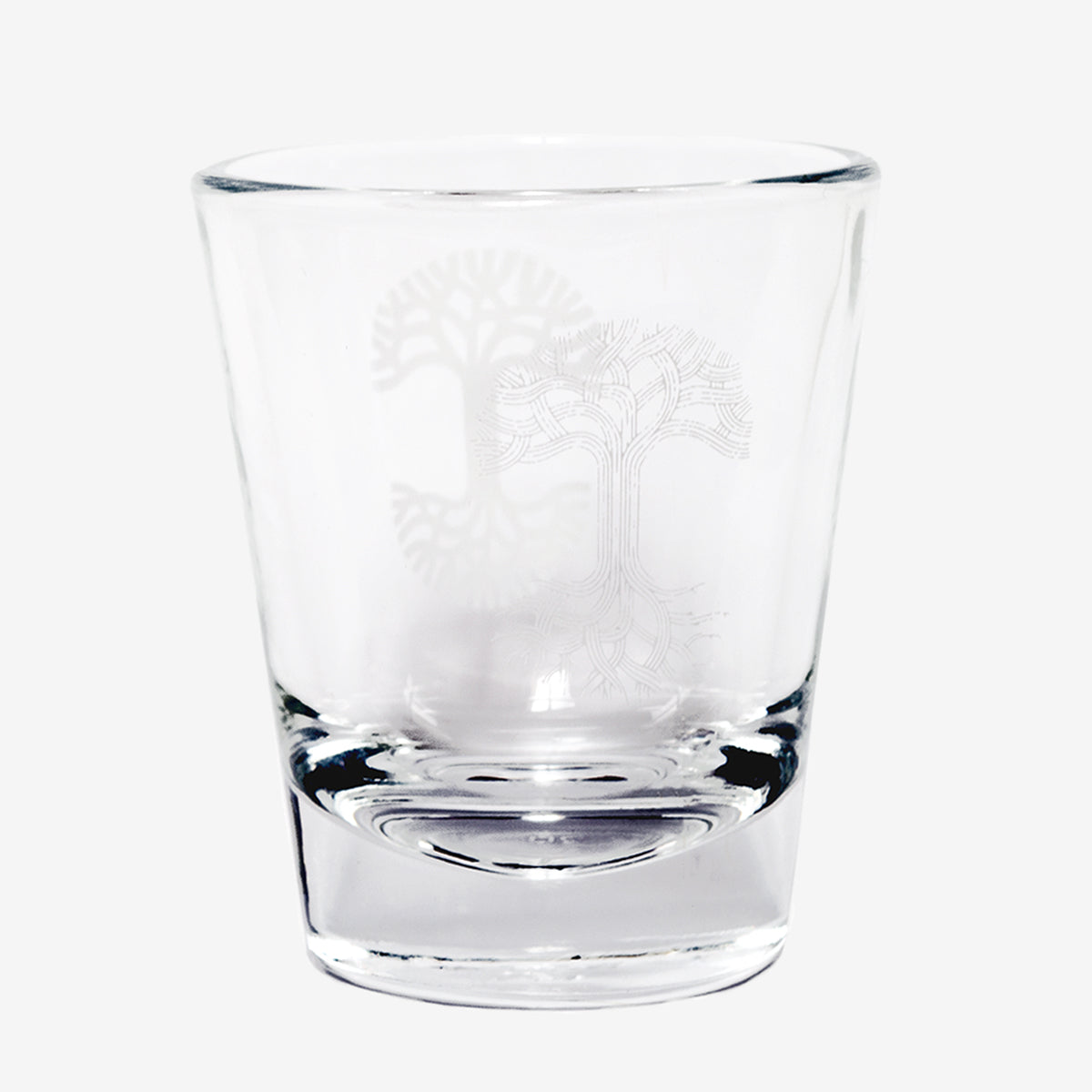 An Oaklandish Trees Shot Glass by Oaklandish is displayed against a plain white background. The clear glass features an etched tree design with intricate, branching roots and branches, forming a symmetrical and detailed pattern. The shot glass possesses a slight shine and reflection, suggesting it is clean and smooth.