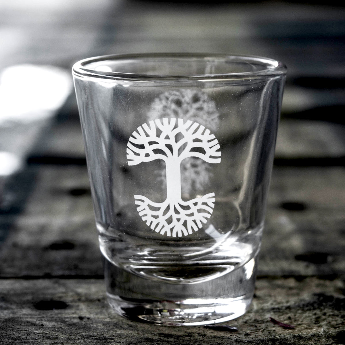 An Oaklandish Trees Shot Glass, featuring a white, stylized tree logo etching on the front, sits on a wooden surface. The blurred background highlights the intricate design of this clear shot glass by Oaklandish.