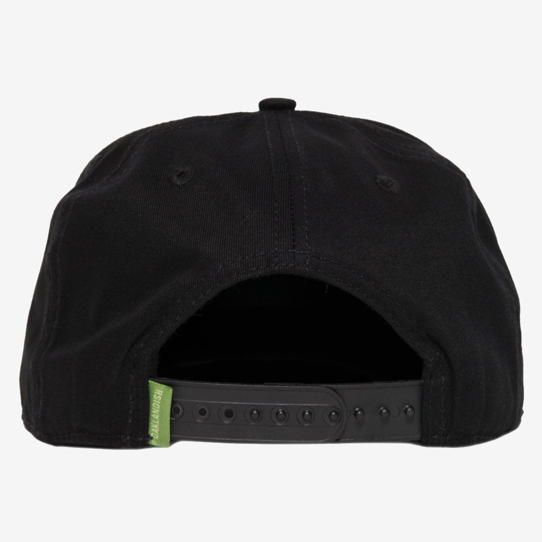The Greenback Snapback by Oaklandish is shown from the back, showcasing a plastic snapback closure with multiple adjustment holes. The black cotton fabric features a small green Oaklandish tag on the left side of the closure. The cap's top button and two ventilation eyelets are visible as well.