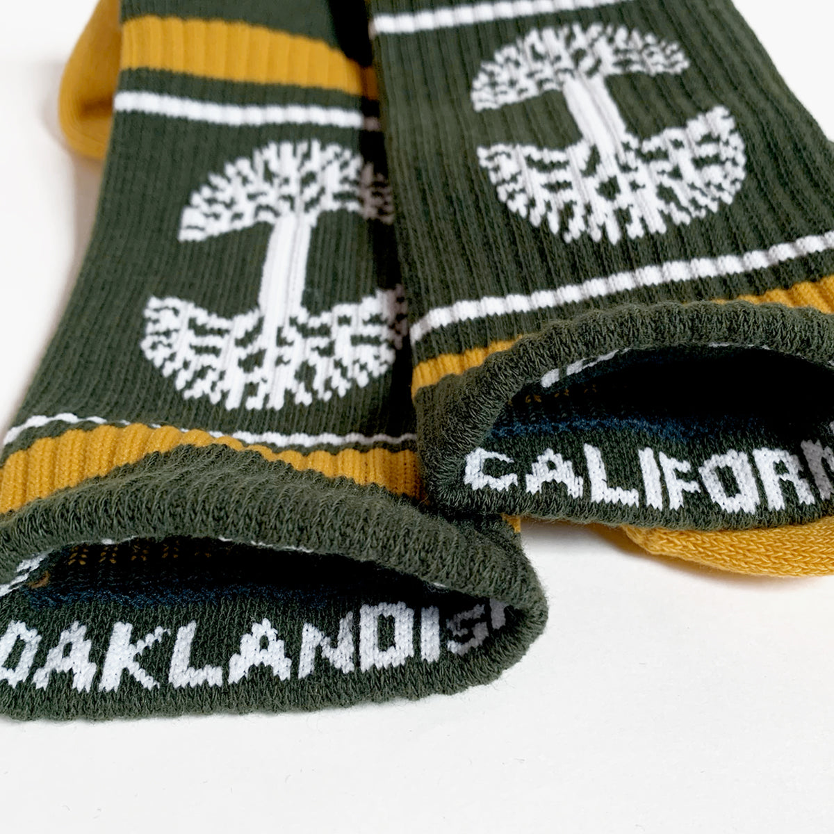 Close-up of two forest green and yellow OAK Knit Crew Socks from Oaklandish, featuring white trees and stripes near the cuffs. The word "CALIFORNIA" is visible on one sock's foot area, and "OAKLANDISH" on the other. These crew socks are displayed on a white surface and are made from cotton, polyester, and spandex.