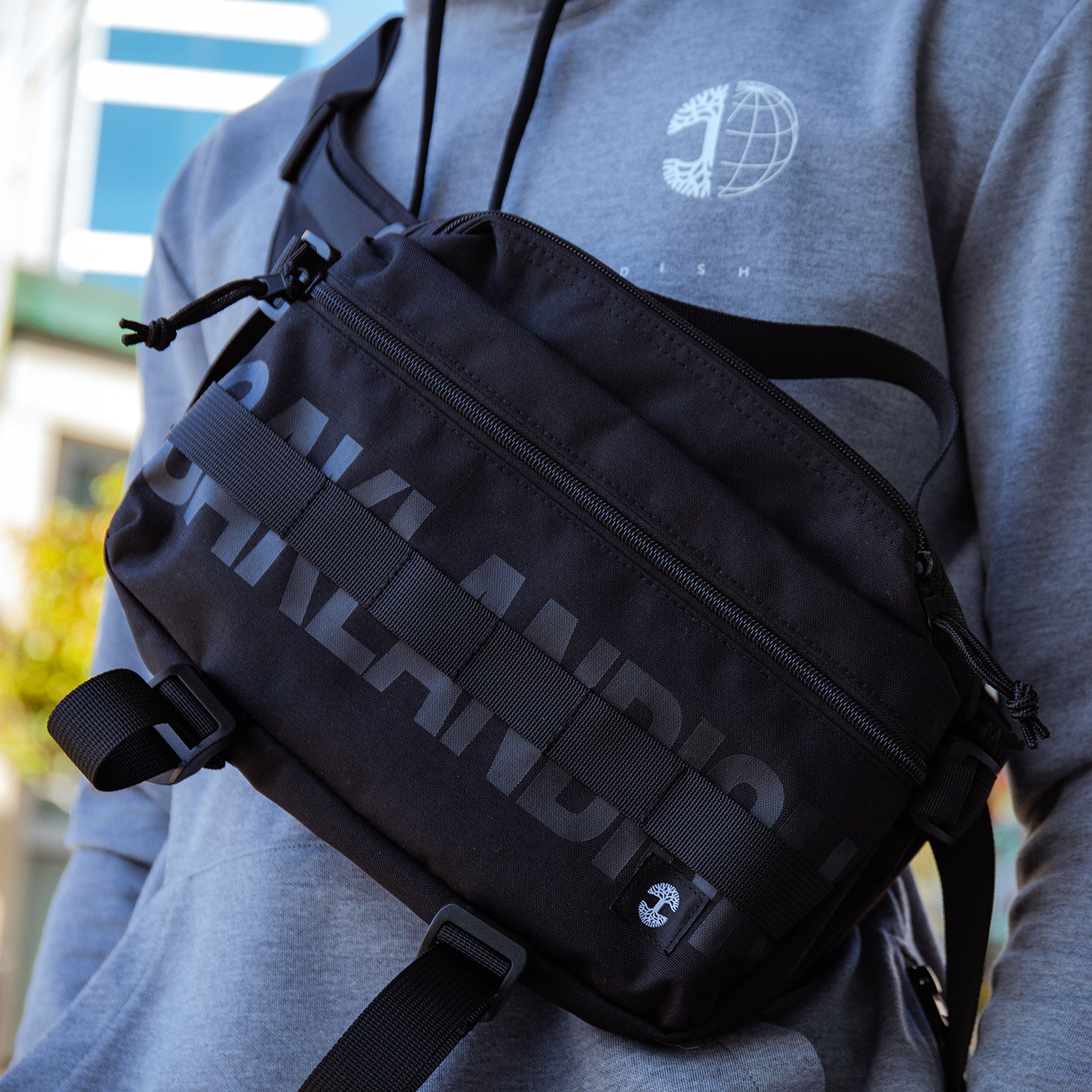 Close-up of person wearing a gray hoodie with a white globe logo and carrying an Oaklandish Hip Bag in black. The unisex bag, from the brand Oaklandish, has two zippered compartments, with the word "OAKLANDI" partially visible in light gray letters. The background shows blurred buildings and trees under sunlight.