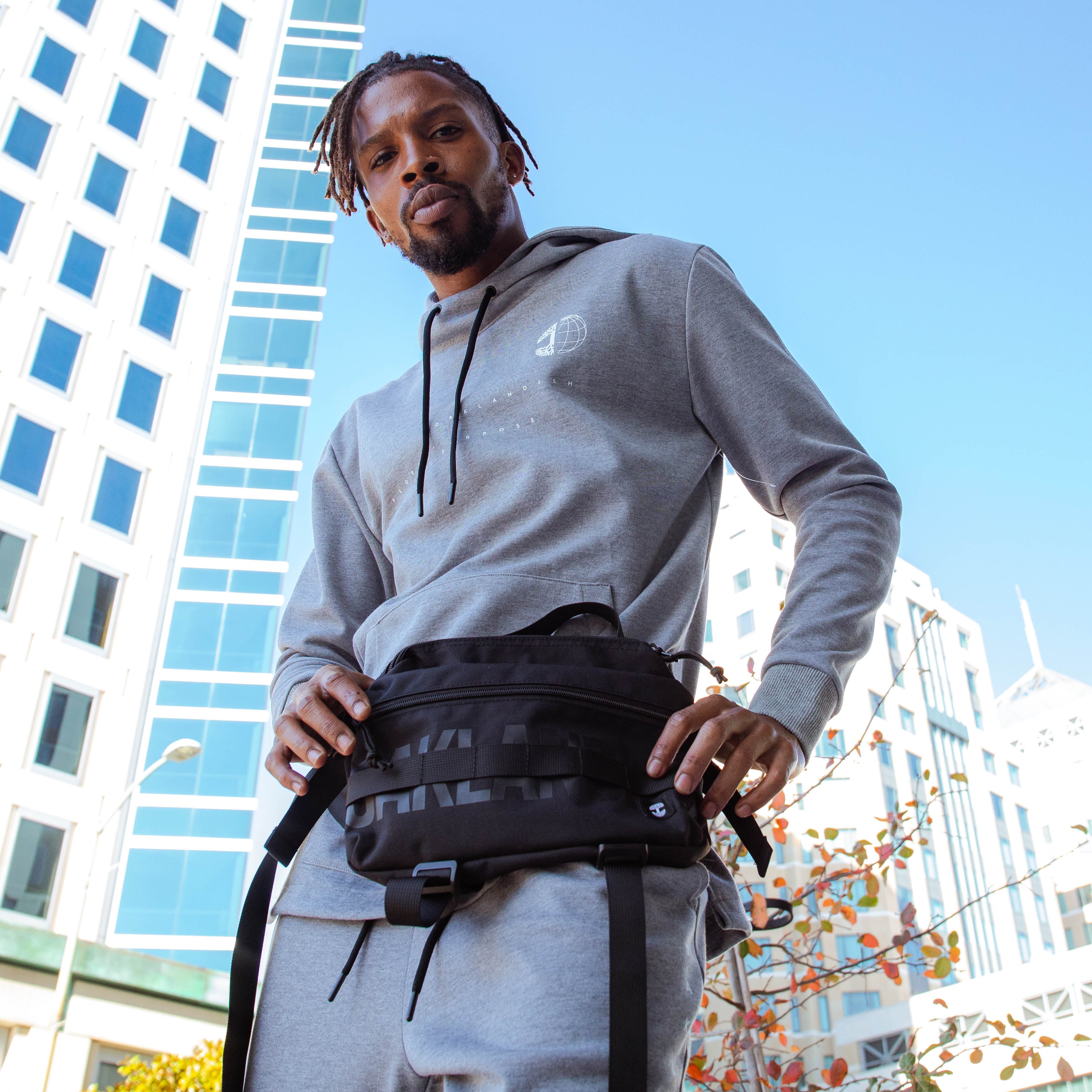 A man stands outdoors in front of modern buildings under a clear blue sky. He is wearing a grey hoodie, matching sweatpants, and an Oaklandish Hip Bag by Oaklandish with large text. His hair is styled in short dreadlocks, and he has a beard. Sparse, leafless branches are visible in the foreground.