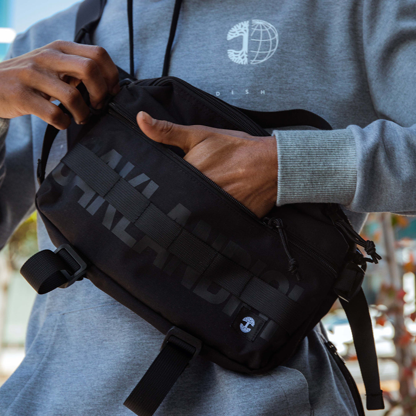 A person wearing a grey hoodie with a globe emblem and the word "SWEDISH" is adjusting their Oaklandish Hip Bag. The black crossbody sling bag, featuring black straps and a visible zipper pocket, complements the sunlit outdoor setting where they stand surrounded by greenery and vibrant bokeh effects in the background.