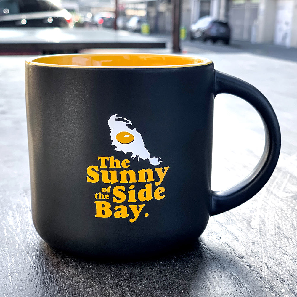 A vibrant Sunny Side Mug by Oaklandish, featuring a glossy yellow interior and sleek black exterior, sits on an outdoor table. The front of this striking Bay mug showcases a graphic of a fried egg and an island shape, along with the phrase "The Sunny Side of the Bay" in cheerful yellow and white text. In the blurry background, cars and a building can be seen.