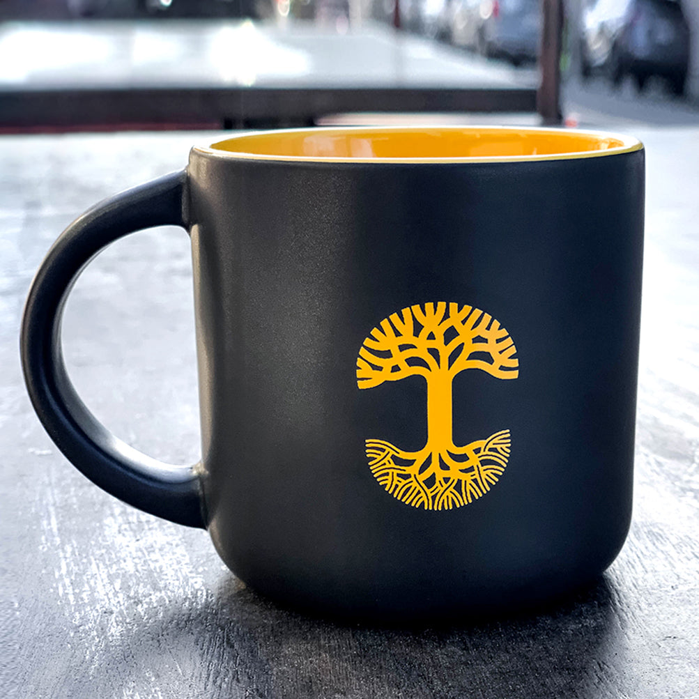 A glossy black Sunny Side Mug by Oaklandish with a vibrant yellow interior sits on a gray table. The mug, also known as the Bay mug, features a striking yellow tree design symbolizing growth and roots. In the blurred background, an outdoor urban setting with indistinct buildings and vehicles can be seen.