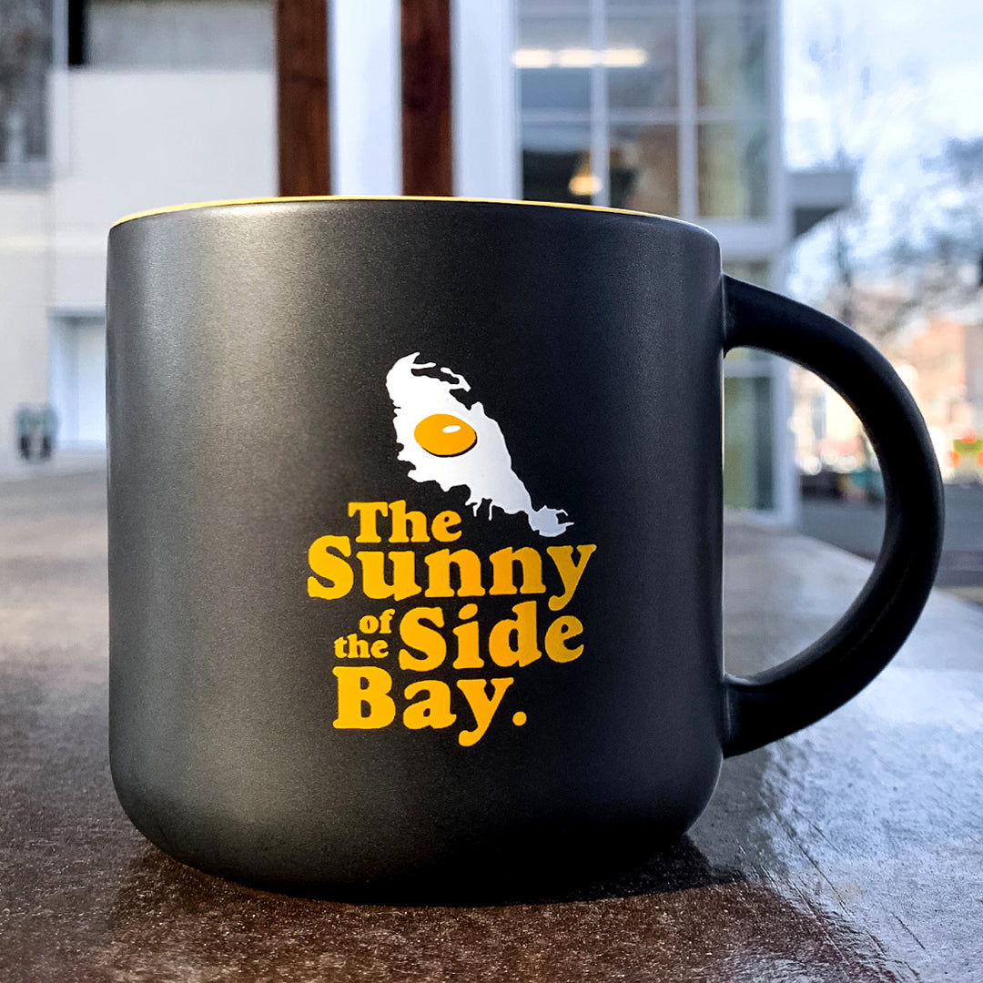 An Oaklandish Sunny Side Mug, with a black stoneware exterior and a glossy yellow interior, rests on a wooden surface. The mug is adorned with yellow text that reads "The Sunny Side of the Bay," complemented by an image of a fried egg shaped like an island. In the background, there's a modern building with large windows and a tree.
