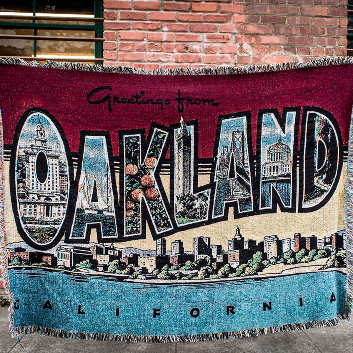 The "Greetings From Oakland Throw" by Oaklandish is a decorative tapestry inspired by vintage postcards. It prominently displays the text "Greetings from Oakland, California," accompanied by vivid illustrations of notable city landmarks such as the skyline, Tribune Tower, City Hall, and scenic waterfront. This unique piece is made from recycled fibers and offers a charming representation of Oakland's iconic scenes.