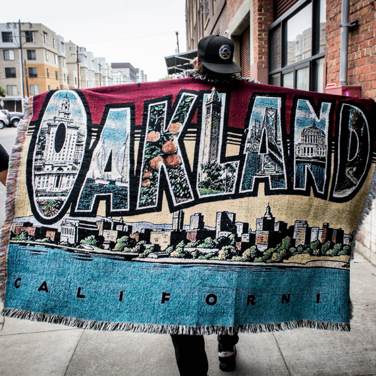 A person wearing a black cap and dark clothing walks down a sidewalk, carrying the large "Greetings From Oakland Throw" by Oaklandish. The tapestry, crafted from recycled fibers, prominently features the word "Oakland" in bold letters and depicts various city landmarks in Oakland, California, set against a vibrant background of blue, tan, and burgundy.