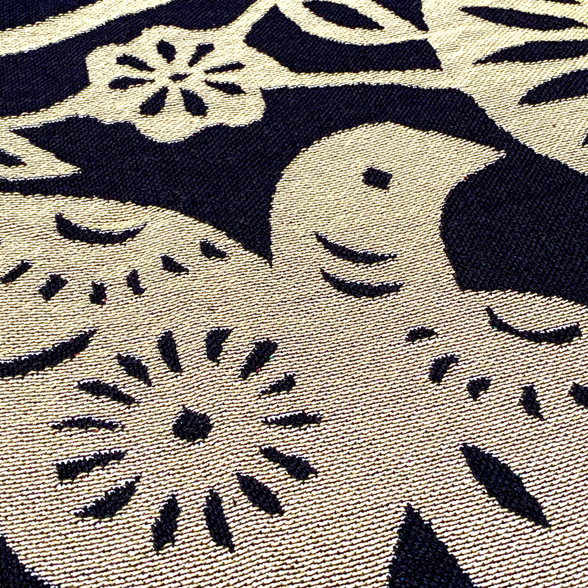 Close-up of the Viva Oakland Throw by Oaklandish, showcasing a fabric design in black and beige tones featuring an intricate bird motif. The bird is surrounded by various floral and leafy patterns, creating a nature-inspired tapestry with bold contrasts. The texture is woven from recycled fibers, adding depth to the detailed surface.