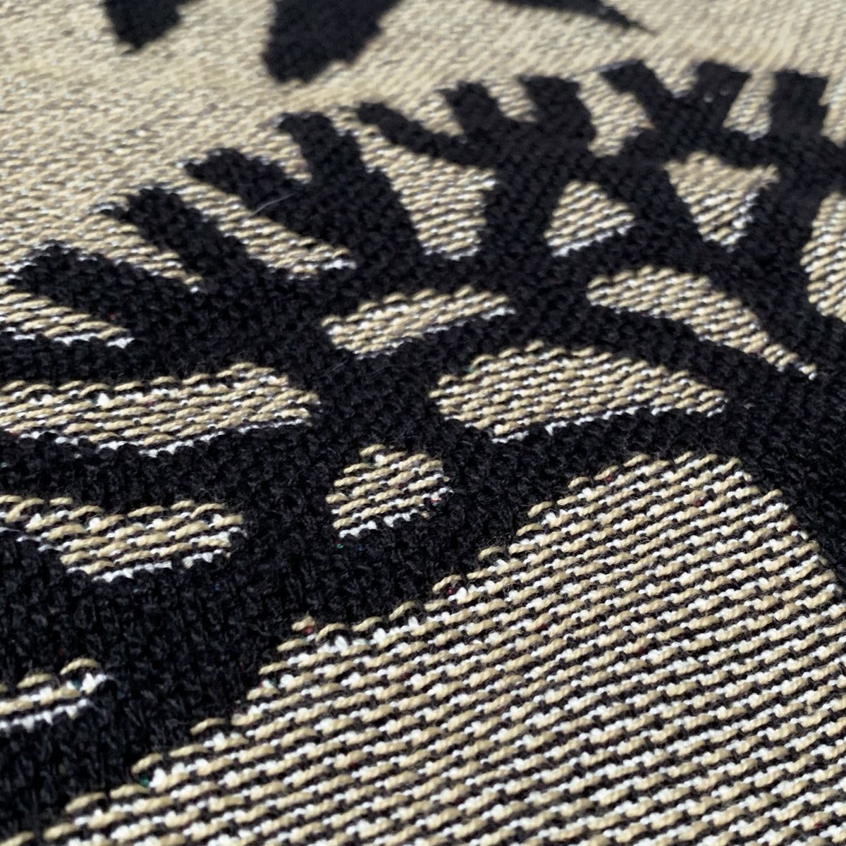 A close-up photo of the Viva Oakland Throw by Oaklandish, featuring a black tree design. The tree is intricately patterned against a beige and off-white background, with visible texture and thread detailing from recycled fibers. The fabric appears tightly knit, displaying clear and sharp contrasts between the colors.