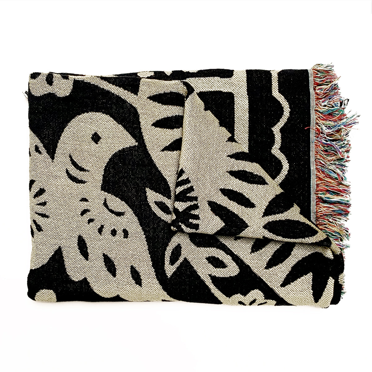 The Viva Oakland Throw by Oaklandish showcases a folded woven design featuring a black and beige bird pattern interspersed with foliage motifs. The edges are adorned with multicolored fringe, adding a touch of vibrant color to the monochrome design. Made from recycled fibers, the fabric appears textured, suggesting a cozy and warm material.