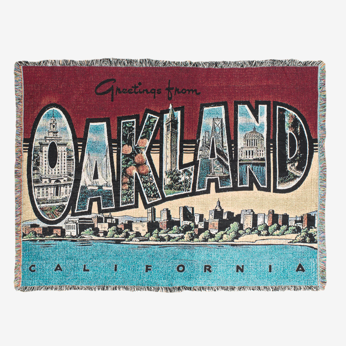 The Greetings From Oakland Throw by Oaklandish is a woven tapestry made from recycled fibers. It features a vintage postcard-inspired design with the text "Greetings from OAKLAND California," where "OAKLAND" contains images of landmarks, buildings, and trees within each letter. The greeting is highlighted against a red background at the top and complemented by a blue background with buildings at the bottom.