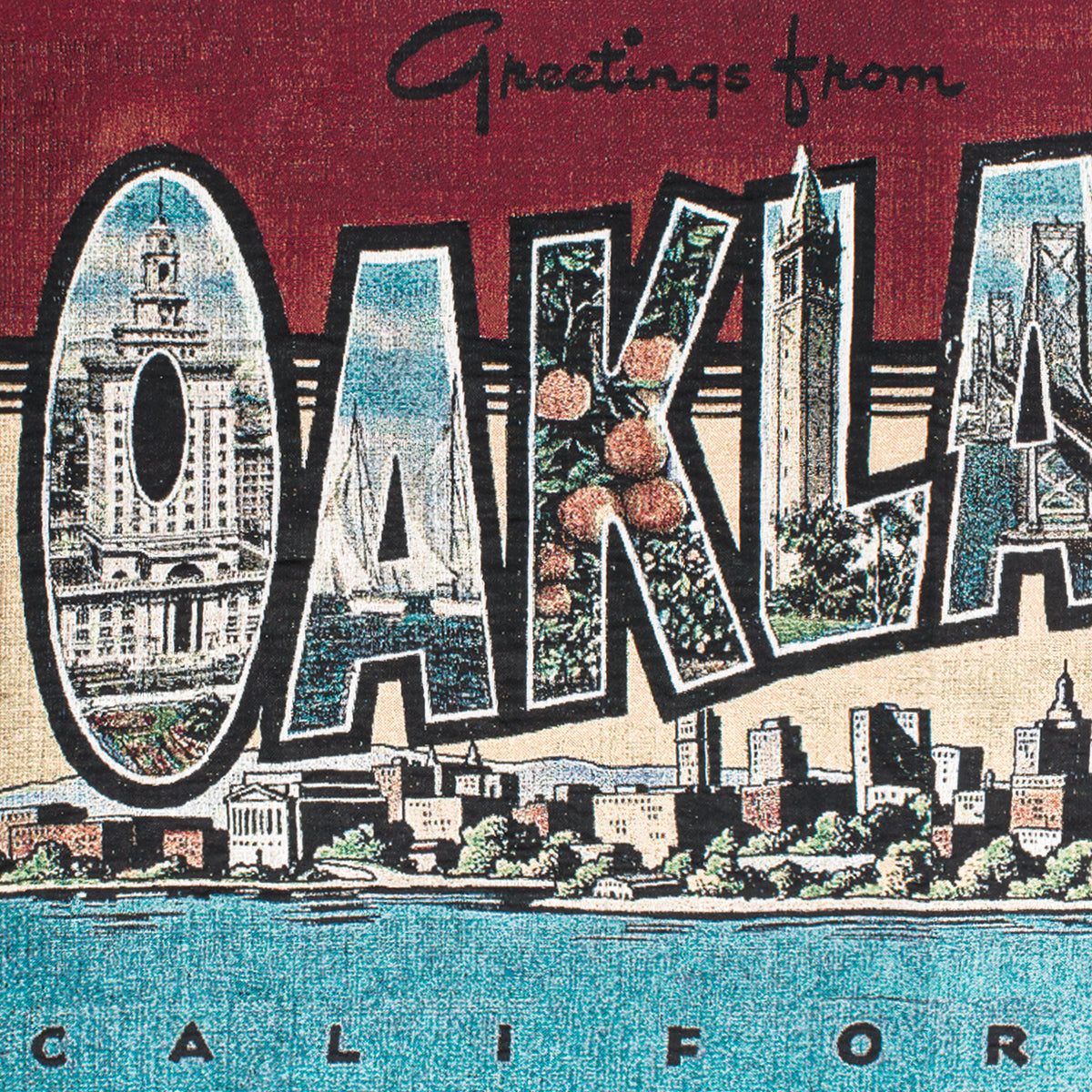 The "Greetings From Oakland Throw" by Oaklandish features a vintage postcard-inspired design with "Greetings from OAKLAND CALIFORNIA" prominently displayed in bold letters. Each letter in "OAKLAND" is adorned with images, including the Tribune Tower, boats, fruit, and the Bay Bridge. Below this text is an illustration of Oakland's skyline complete with trees and buildings set against a blue water foreground. This throw is made from recycled fibers.