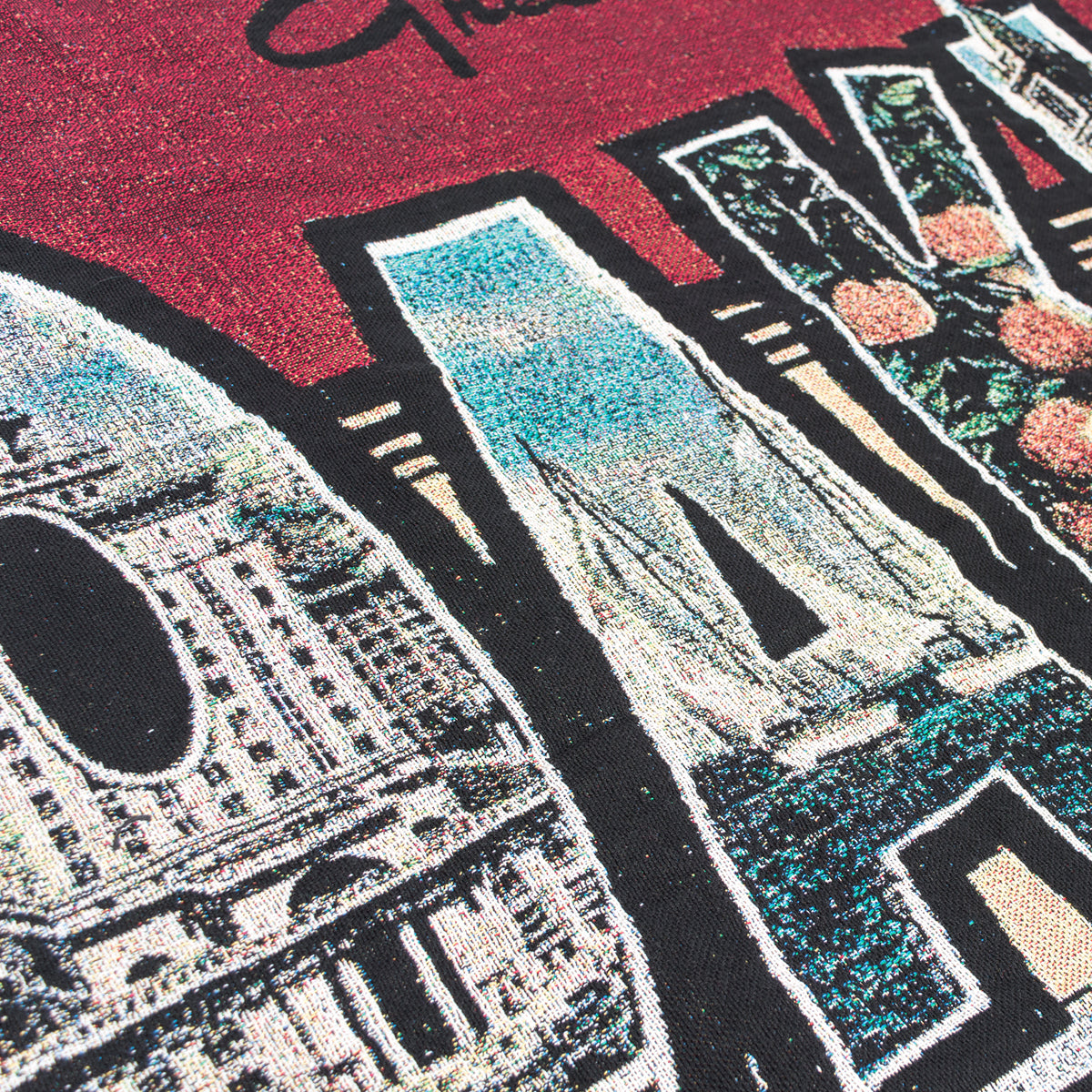 Close-up of the Greetings From Oakland Throw by Oaklandish, featuring a graphic design on a burgundy fabric with the word "SAN" in large, stylized letters. Each letter is filled with a vintage postcard-inspired collage of colorful images depicting scenes like architecture, landscapes, and flowers. The vibrant design has a textured appearance and uses recycled fibers.
