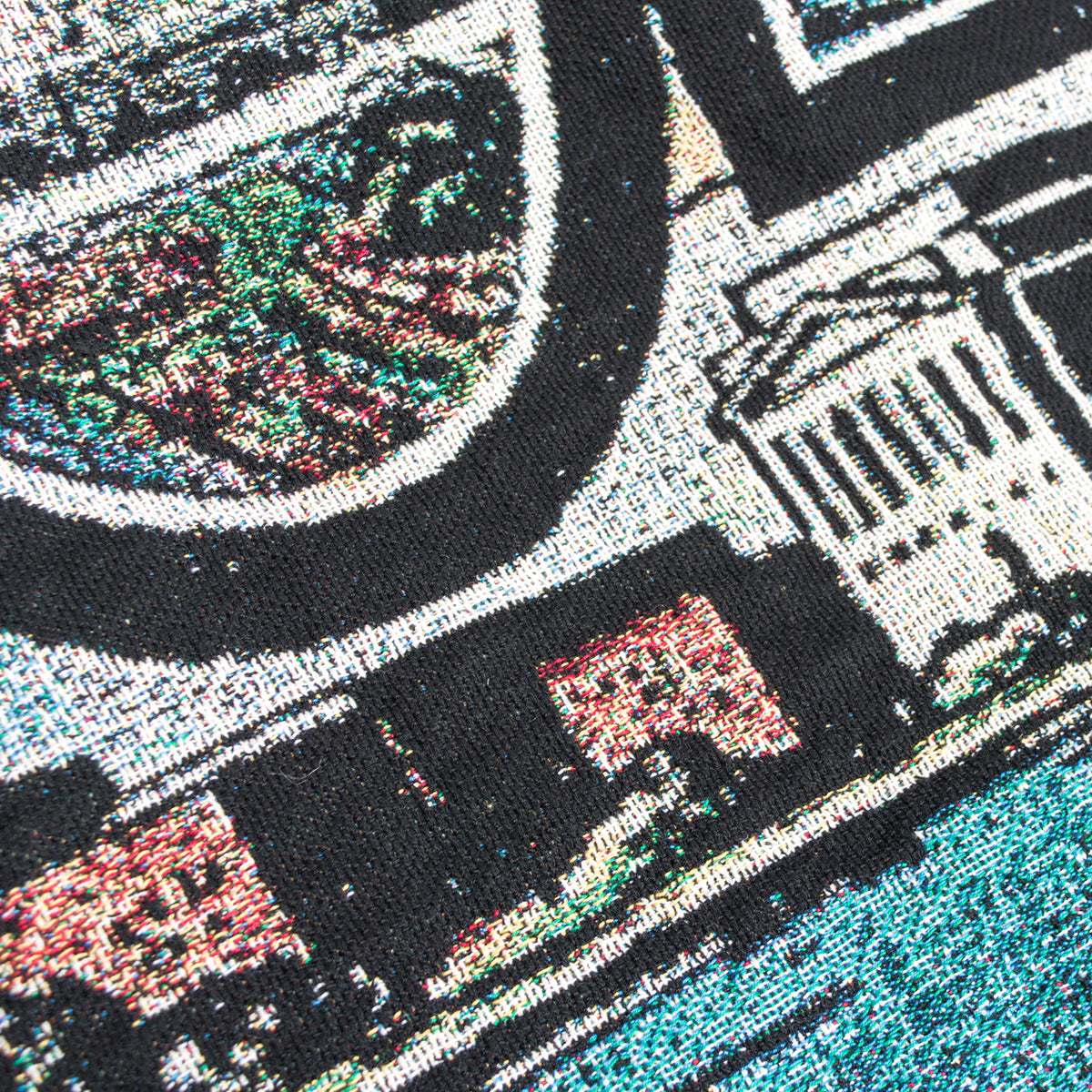 A close-up view of the Greetings From Oakland Throw by Oaklandish, woven with intricate cityscape patterns reminiscent of a vintage postcard. The design includes various buildings, greenery, and a body of water, rendered in bright colors with black outlines. The detailed fabric texture highlights the recycled fibers used in the weave.