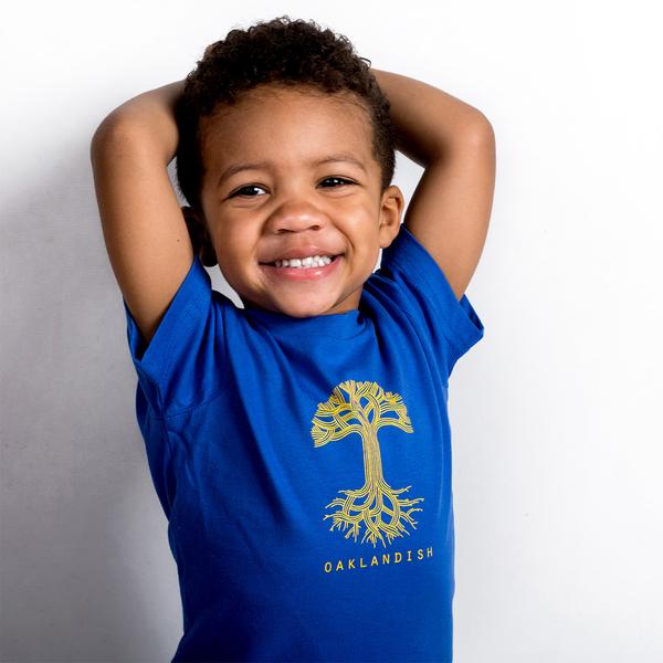 A young child with curly hair is smiling widely, wearing a bright royal blue "Toddler Oaklandish Classic Logo Tee" by Oaklandish. The 100% cotton shirt features a yellow tree design and the word "OAKLANDISH" printed below the graphic. The child has their hands behind their head, posing in front of a white background.