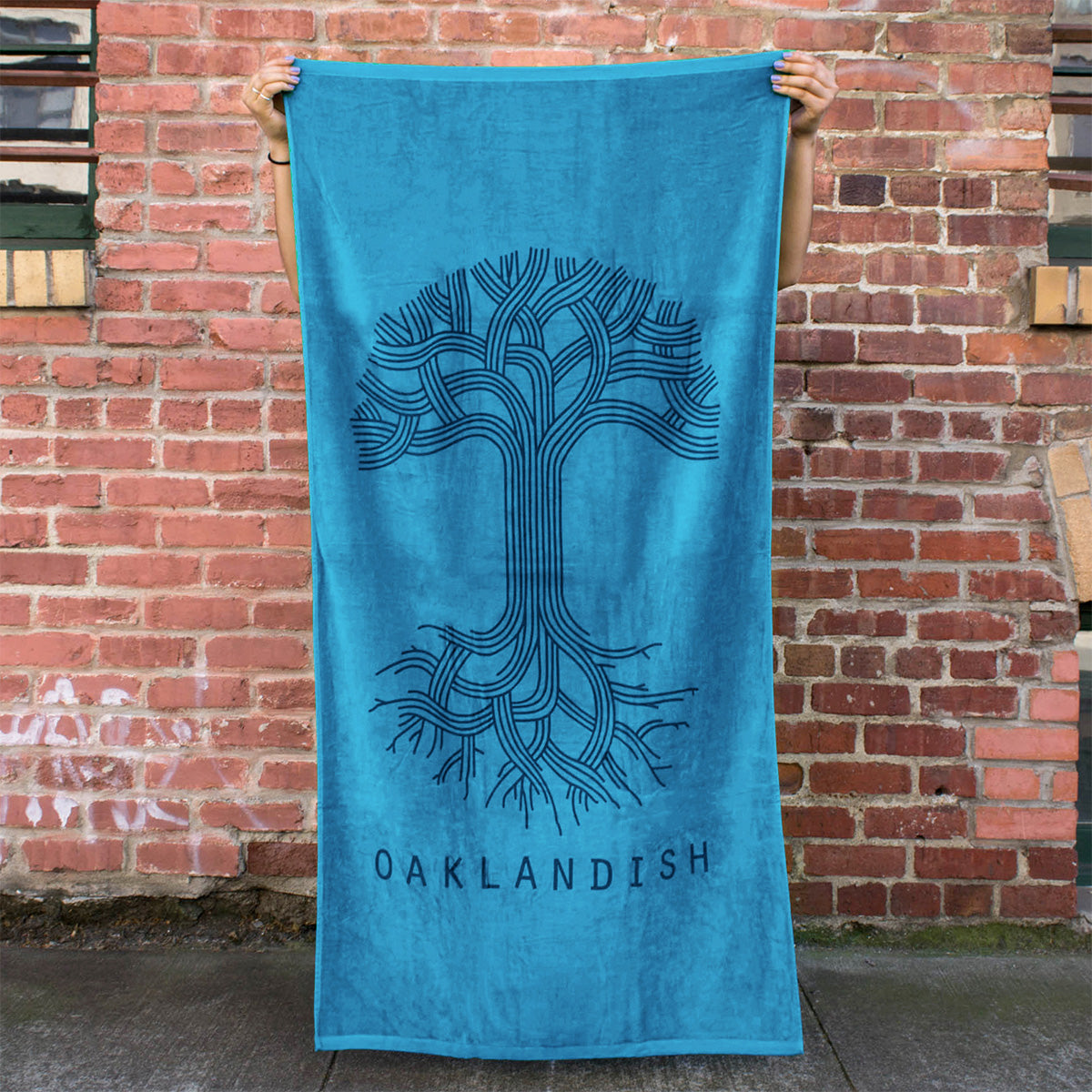 A person is holding up an oversized Classic Logo Beach Towel by Oaklandish, made of cotton terry velour in turquoise. The towel features a stylish dark tone-on-tone print of a stylized tree with intricate branches and roots, with the word "OAKLANDISH" printed at the bottom. The background showcases a rustic brick wall adorned with windows and visible graffiti.