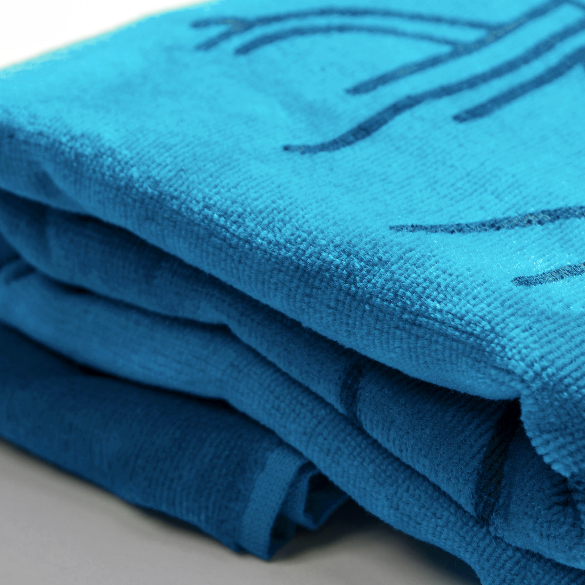 Close-up of a stack of folded, bright blue Classic Logo Beach Towels by Oaklandish, featuring a decorative stitched pattern. The texture of these oversized plush cotton terry velour towels appears soft and lush. The neatly arranged towels rest on a smooth, light-colored surface.