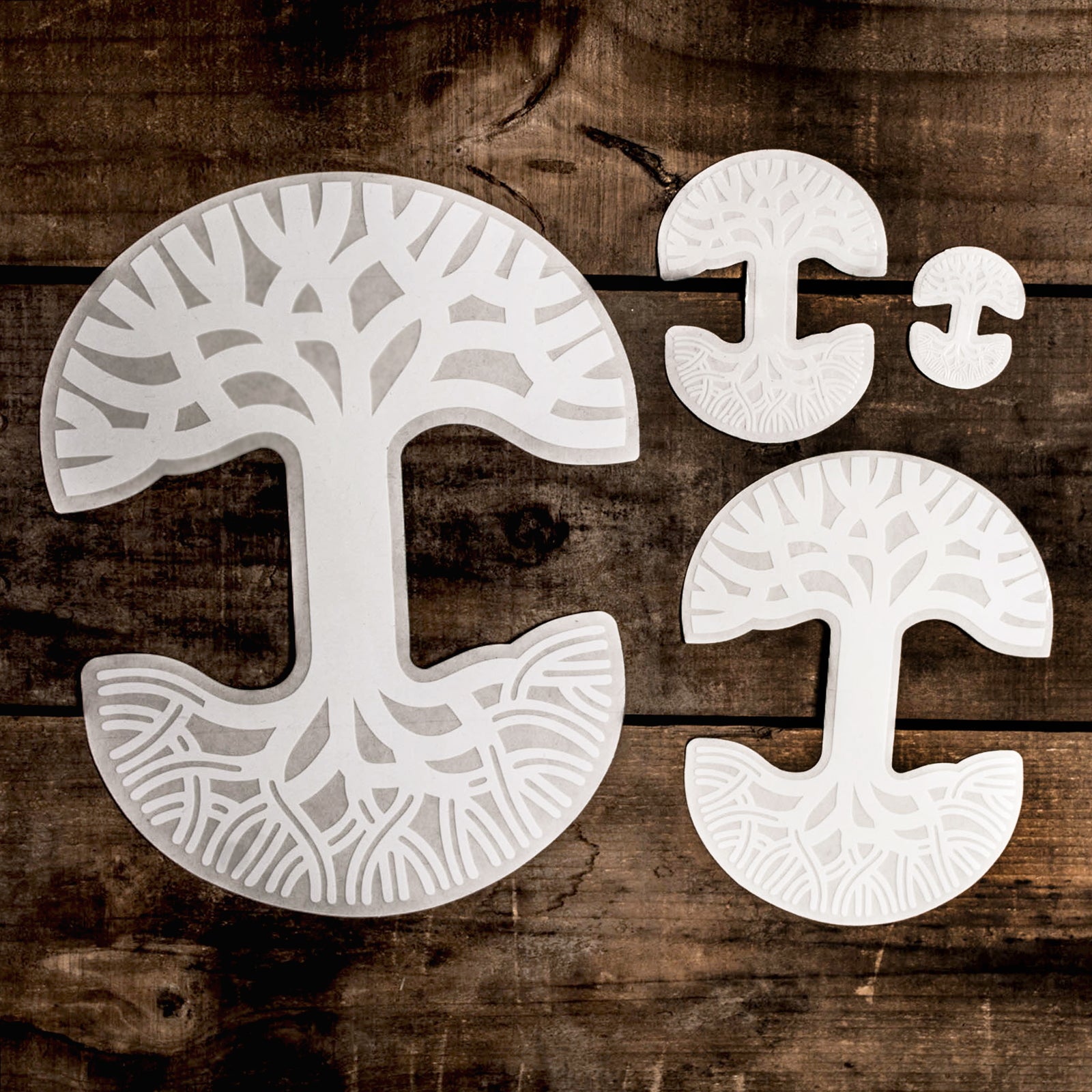 Four Oaklandish Decal 3X4.5 stickers by Oaklandish, featuring intricate tree-shaped designs with branching roots and canopy patterns, are displayed on a wooden surface. These durable outdoors-friendly decals vary in size, with the largest on the left, a medium-sized one on the right, and two smaller ones on the top right.